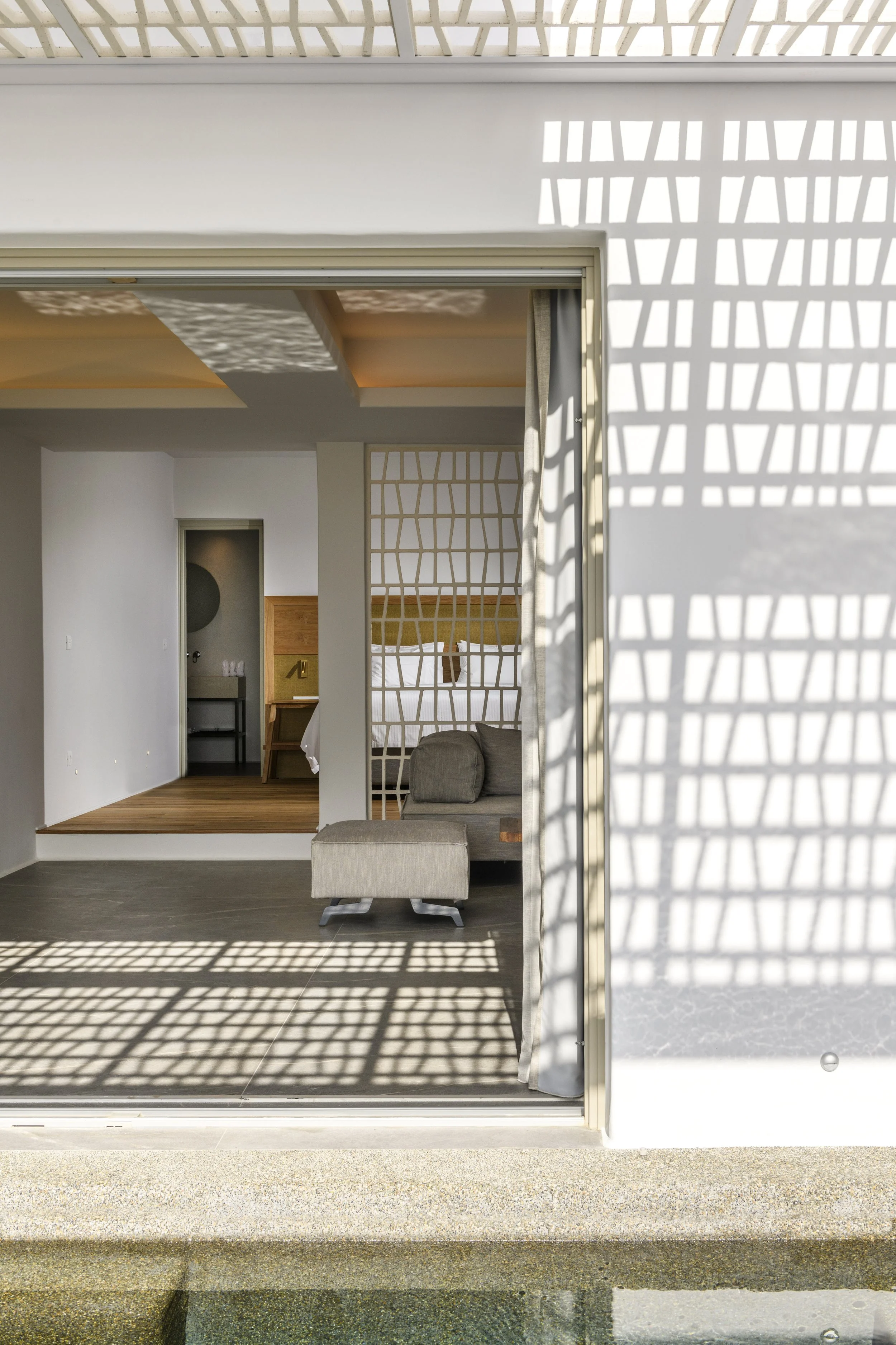 Sunlight casting a shadow pattern of the railing on the white wall and glass sliding door of a modern house. Through the door, part of a living room with a sofa and a bedroom with a bed are visible.