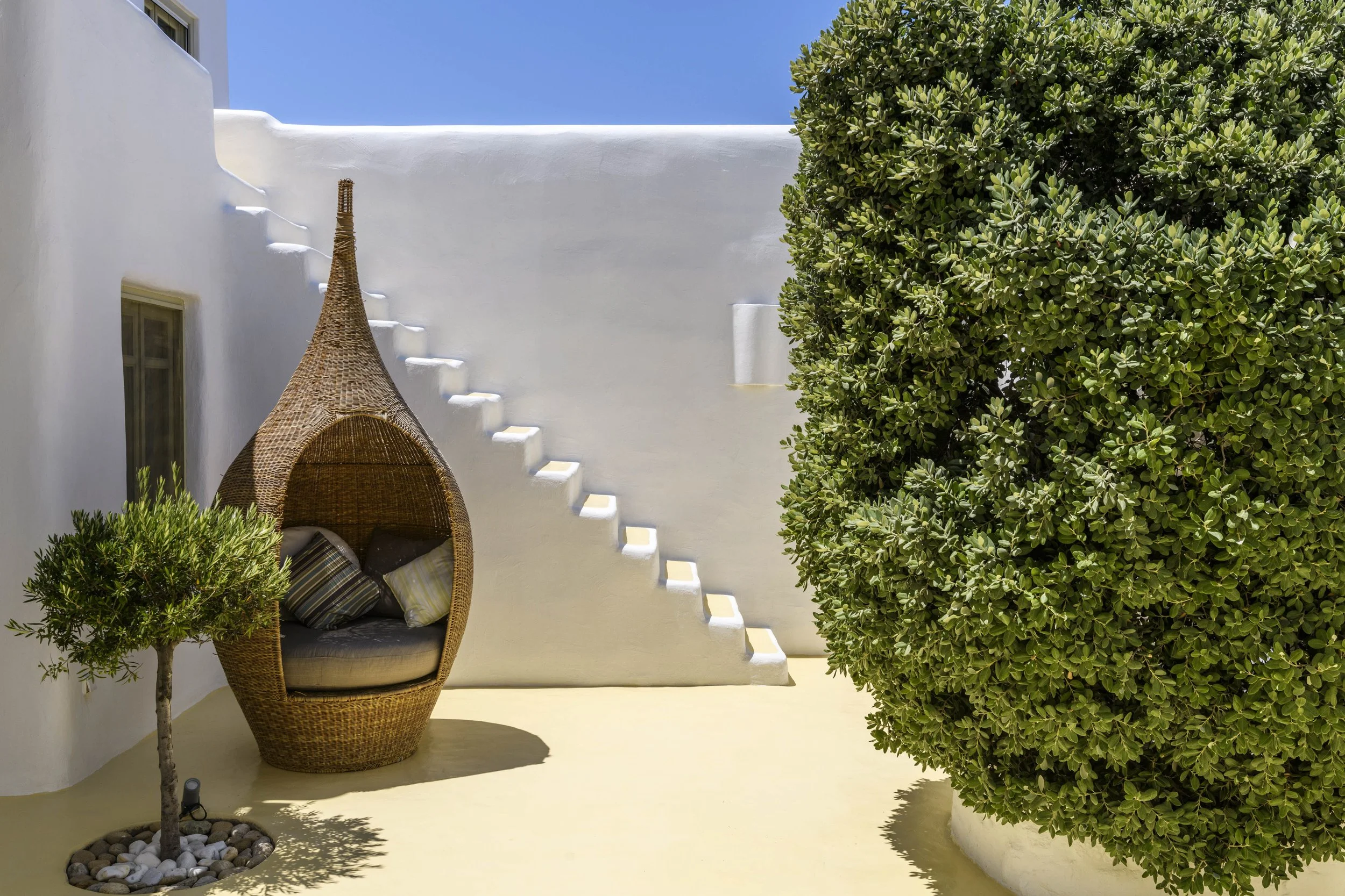 A white stucco wall with stairs, a wicker egg-shaped chair with cushions, a small potted tree with rocks, and a large green bush under a blue sky.