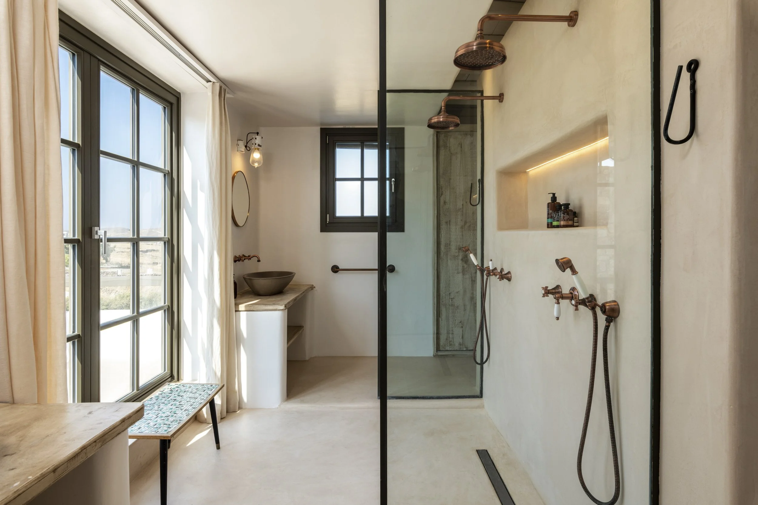 Modern bathroom with large windows, a walk-in shower with copper showerheads, and a stone basin sink on a marble countertop.