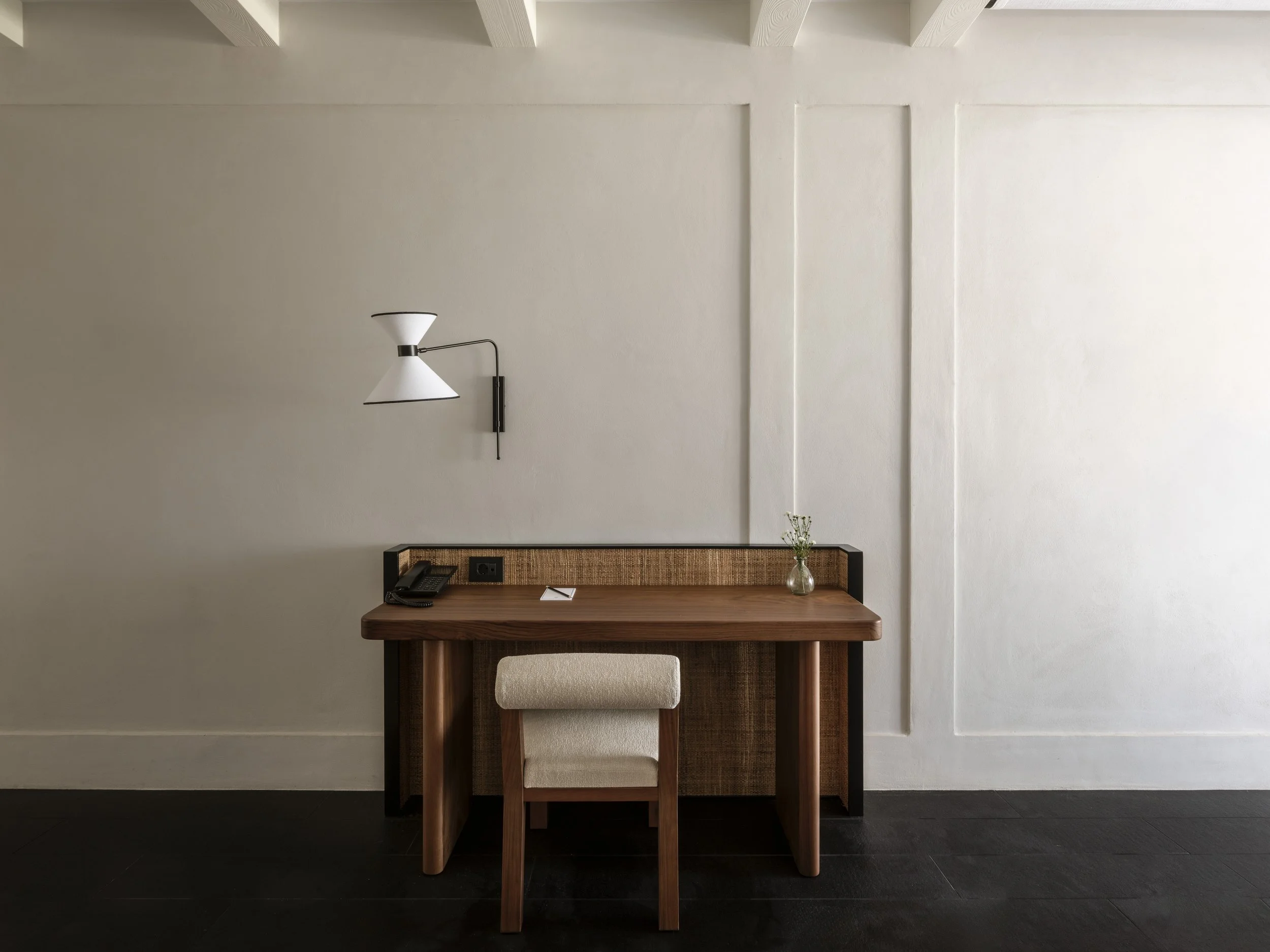 A minimalist interior with a white wall, a hanging white and black wall lamp, a wooden console table, a beige upholstered chair, a small glass vase with white flowers, and some papers and a black remote on the table.