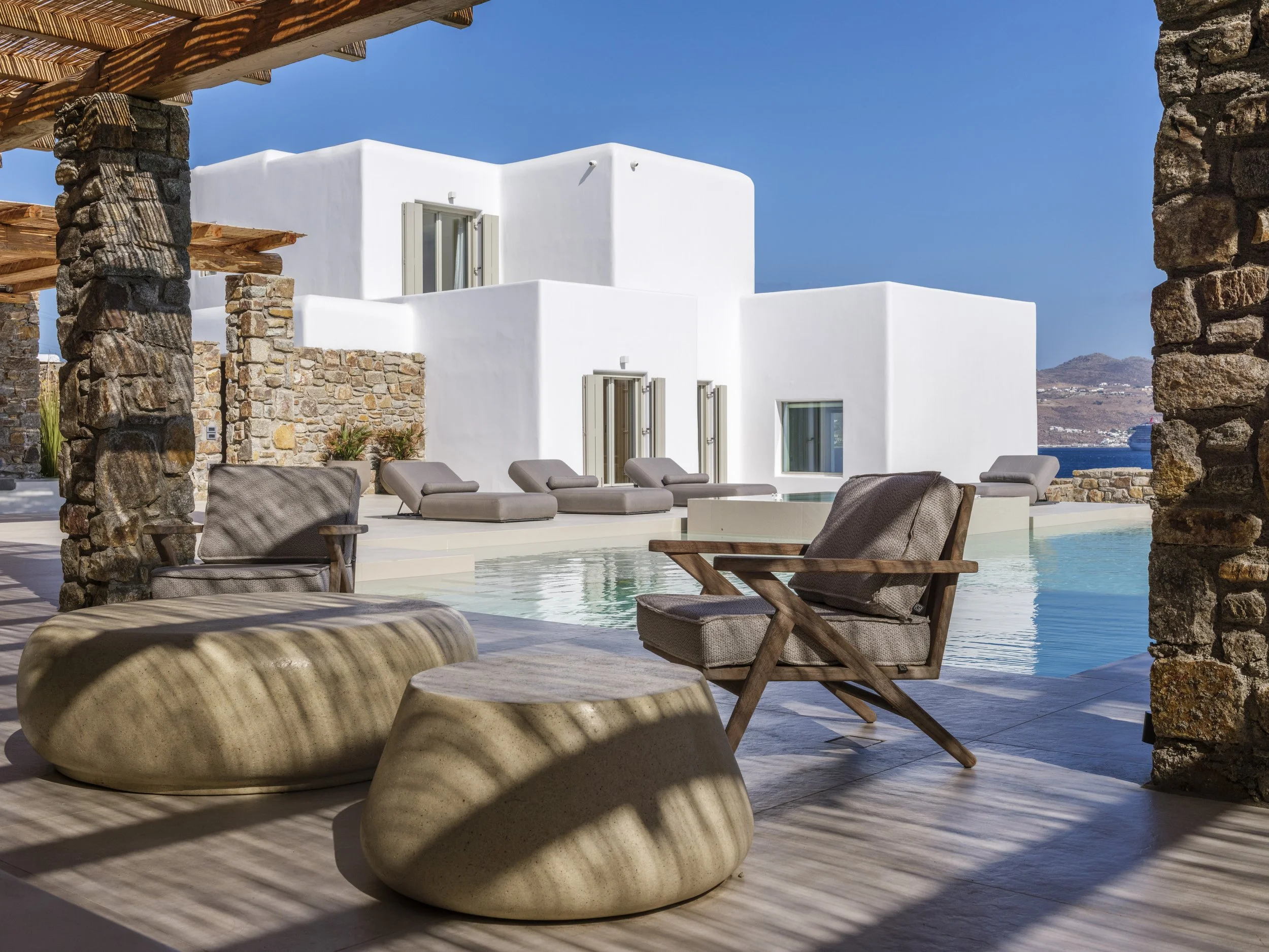 Outdoor seating area with beige stone stools, lounge chairs, and a swimming pool, surrounded by stone columns and white contemporary buildings under a clear blue sky.
