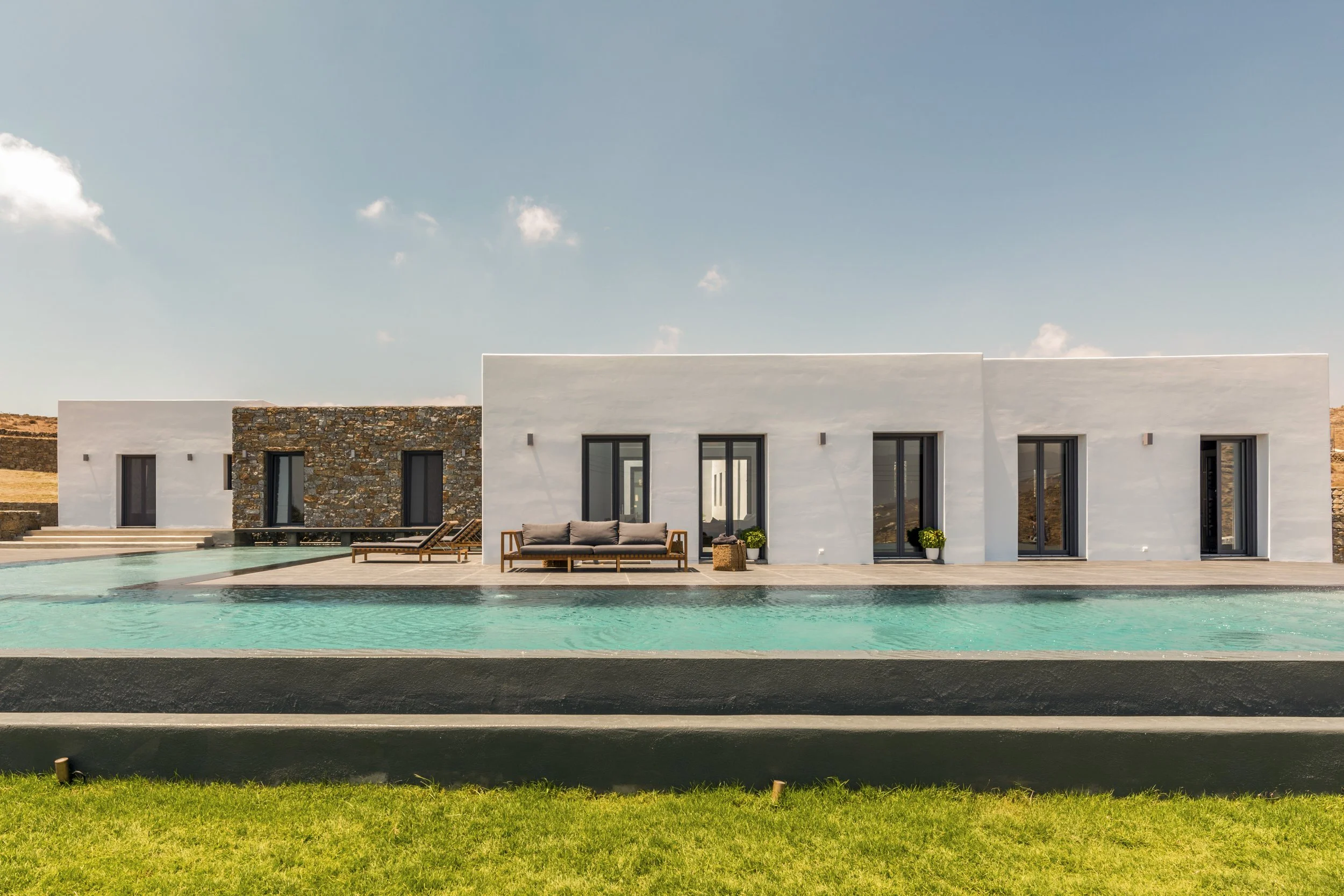 Modern Cycladic Villa located in Mykonos  with large glass doors, a stone accent wall, an outdoor pool, and minimalist outdoor seating, with a backdrop of mountains and a partly cloudy sky.