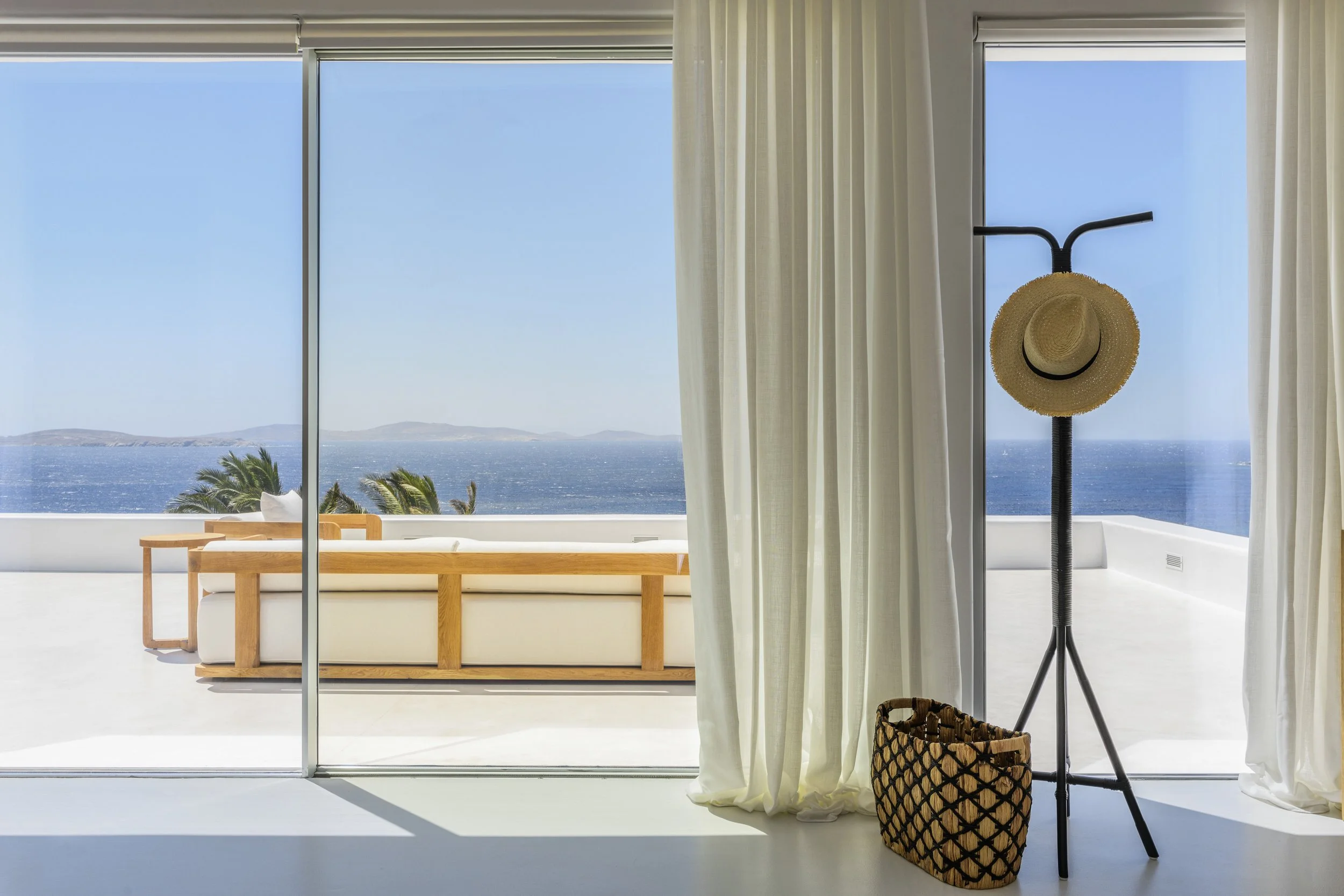 Room with large glass sliding door revealing a view of the ocean and distant hills, with white curtains and a hat on a coat stand near the door, a woven basket nearby.
