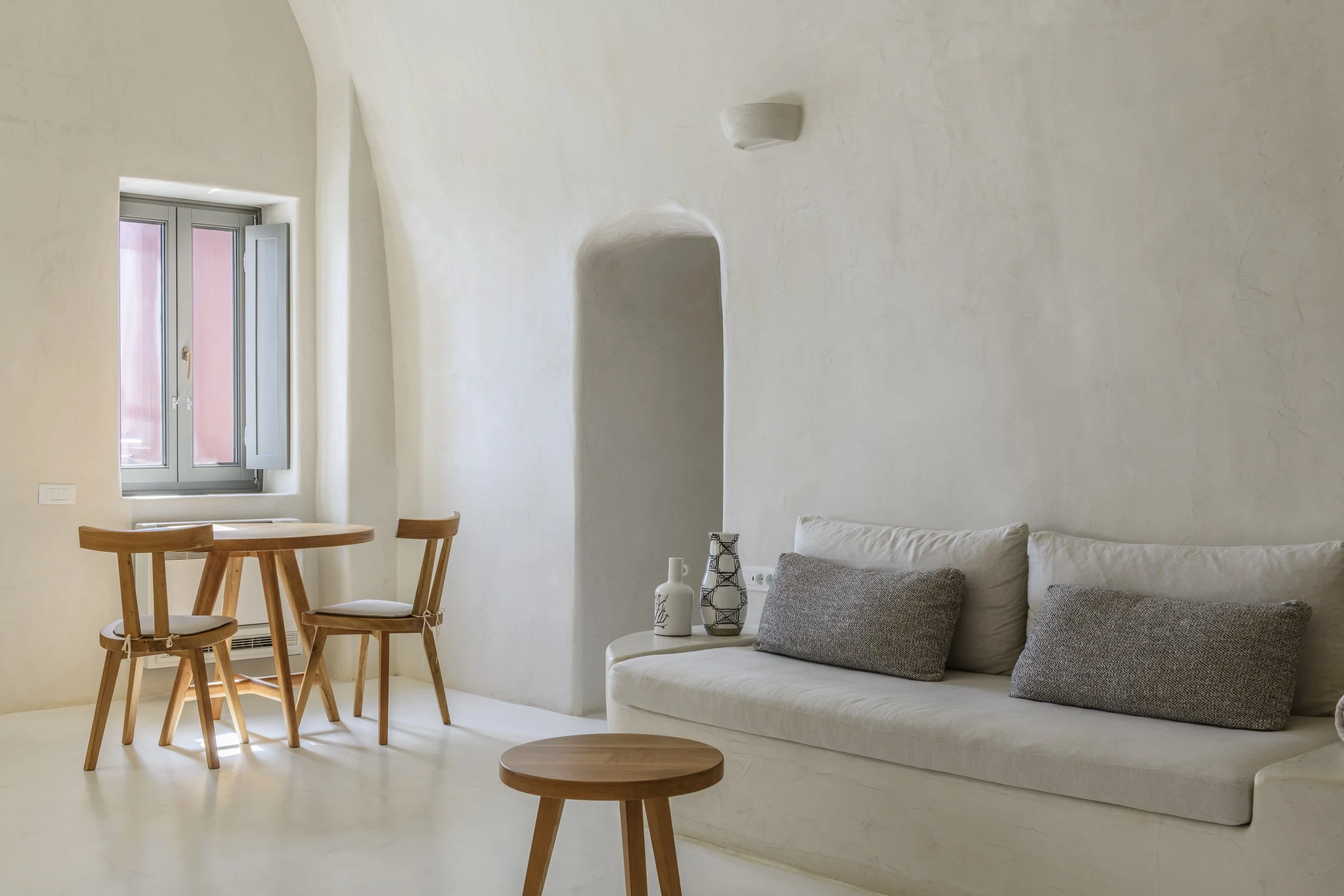 A minimalist living room with white walls, a white sofa with gray pillows, a small white coffee table with decorative vases, a wooden round side table, and a wooden dining table with matching chairs near a window with closed shutters.