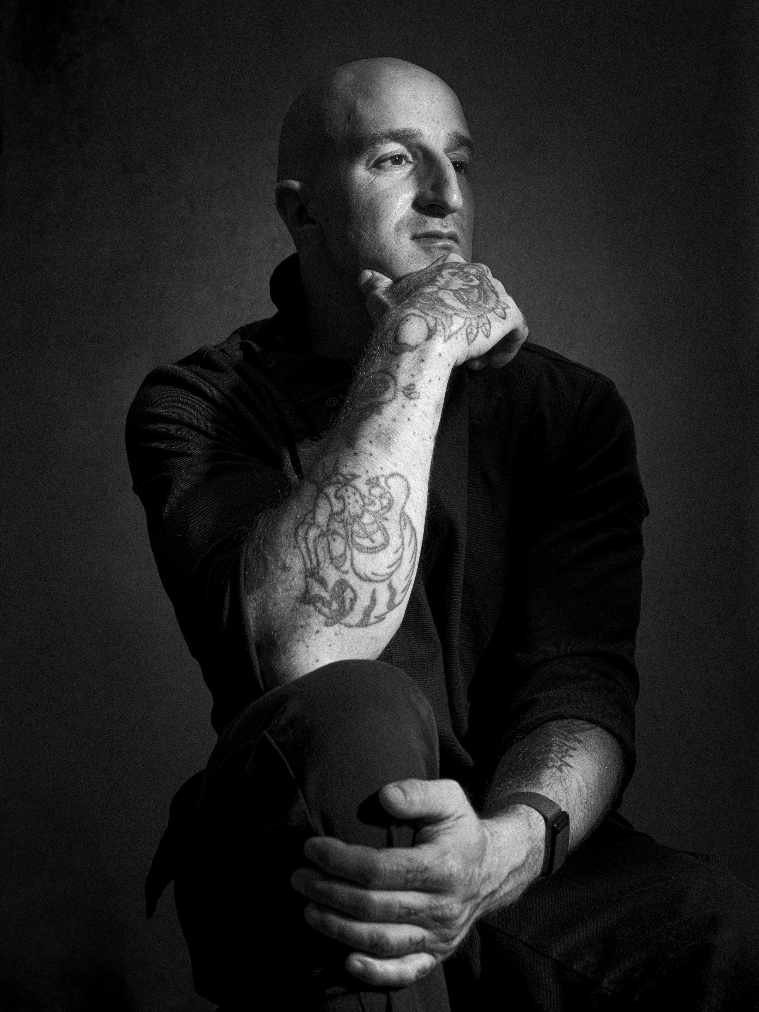 A black and white portrait of a bald man with tattoos on his left arm, resting his chin on his hand, and looking thoughtfully to the side.