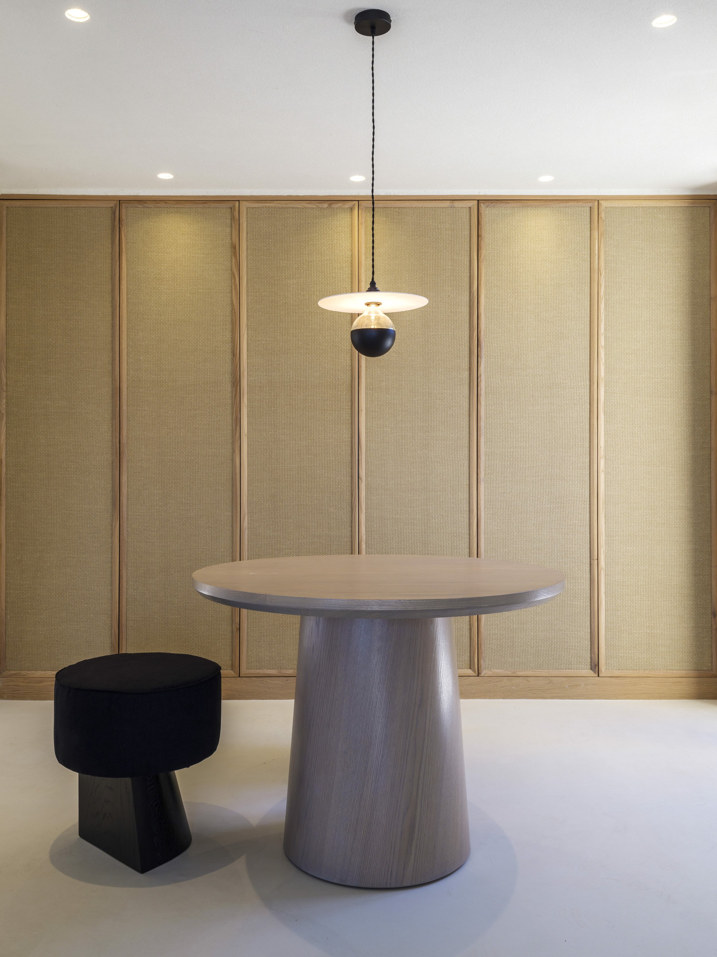 A modern interior space featuring a round table with a textured, cone-shaped base, a black upholstered stool, and a hanging pendant light with a black sphere on a white disc. The background includes a wall with vertical wooden frames and woven panels