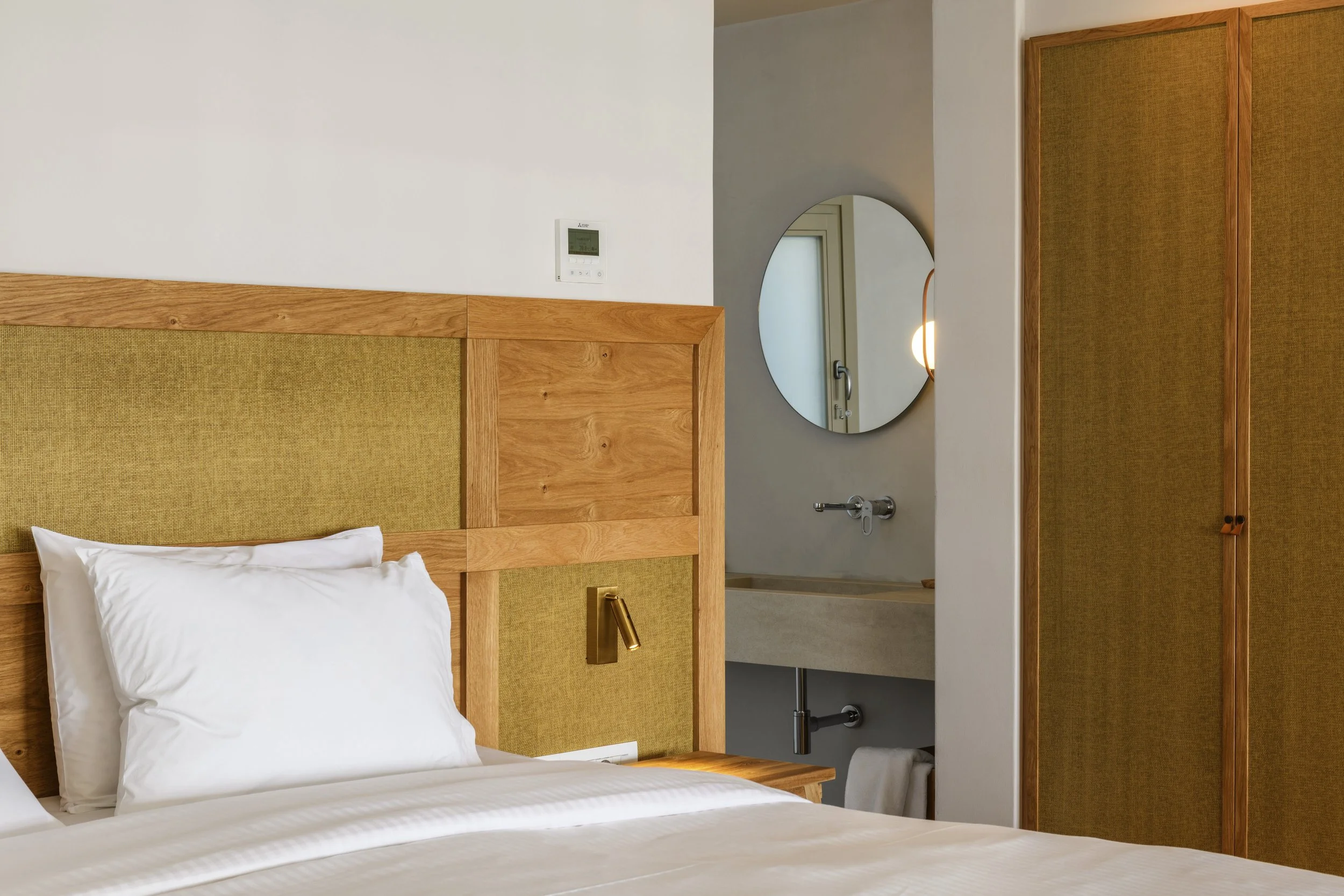 A hotel room with a white bed, a wooden headboard with beige fabric panels, a gold reading lamp, a wall-mounted thermostat, a round mirror, and a view of a bathroom with a towel rack and door.
