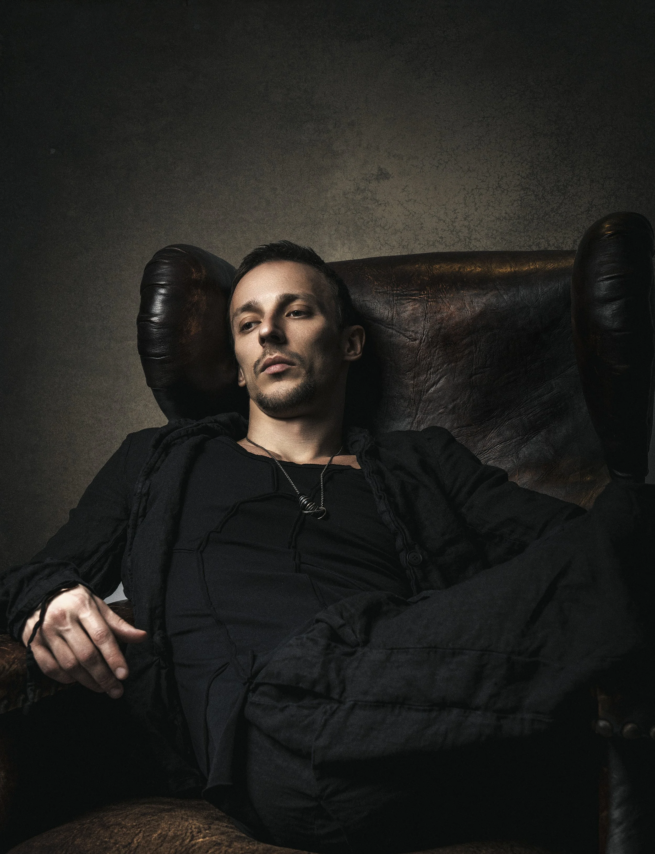 A young man with dark hair and a goatee, wearing a black jacket, is reclining in a large, dark leather armchair. His head rests against the chair's high back, and his left hand is resting on his lap. The background is a plain, dark wall.