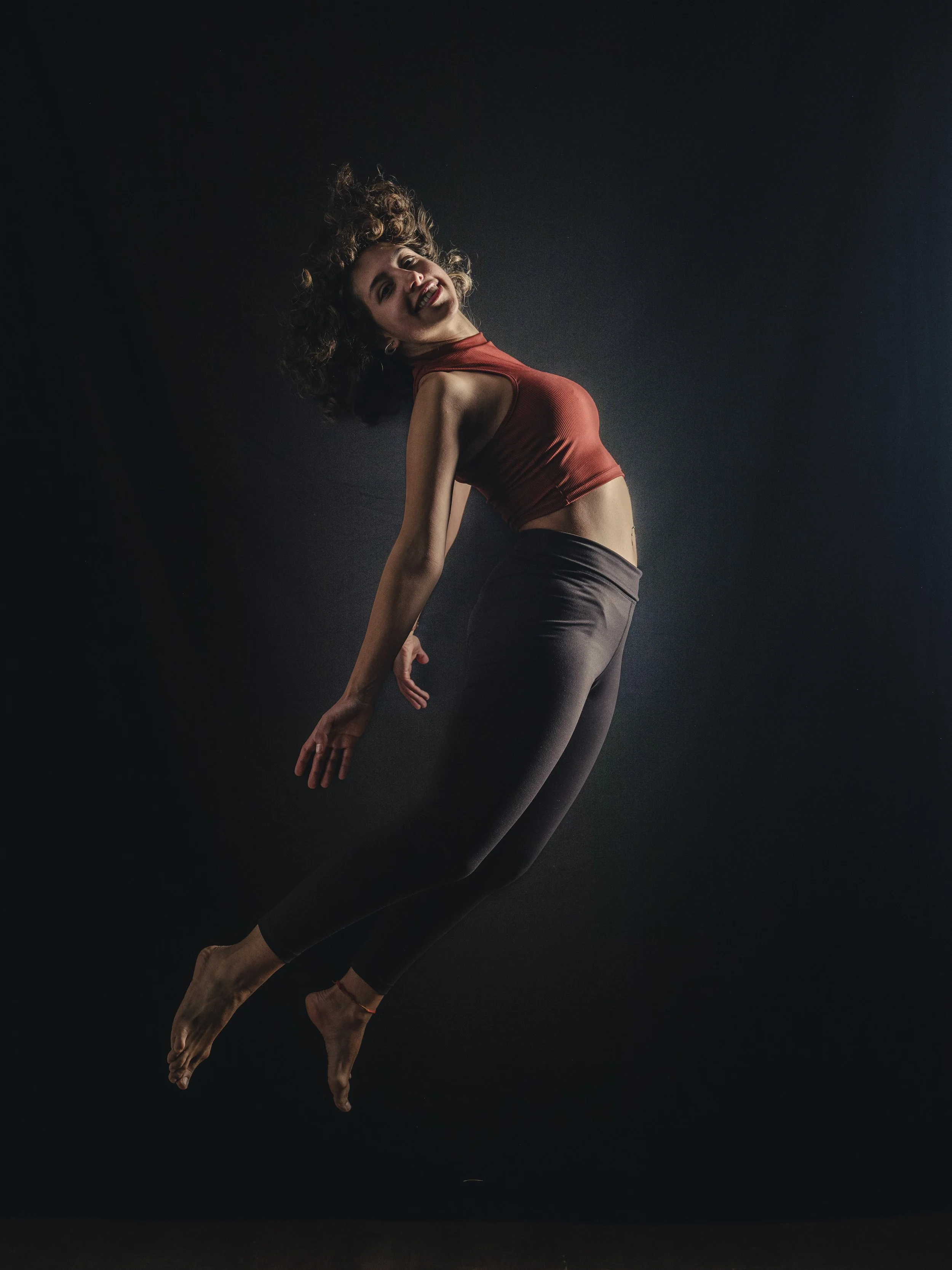 A woman with curly hair, wearing a red sleeveless top and black leggings, poses against a dark background, smiling and arching her back slightly.