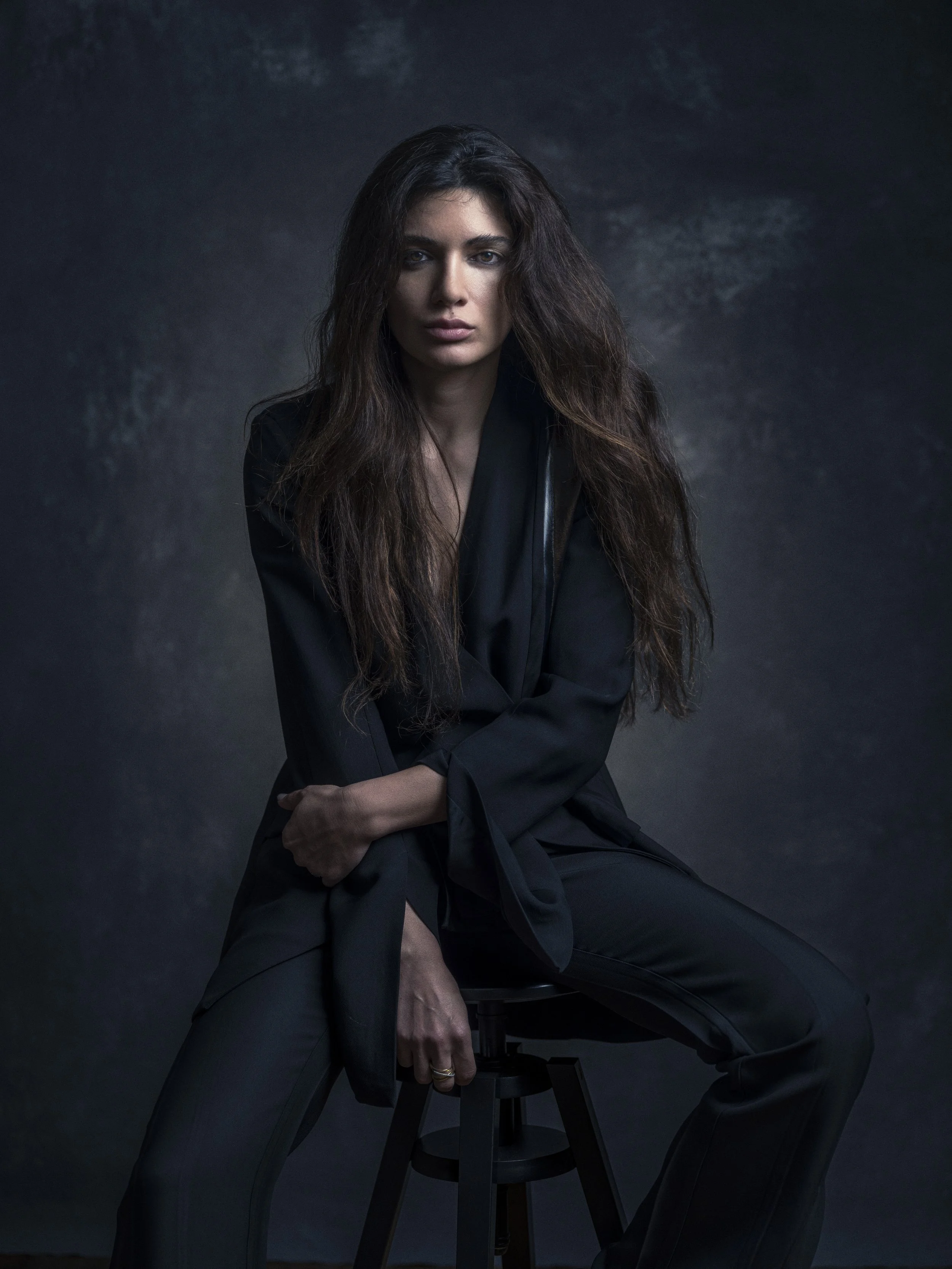 A woman with long, dark, voluminous hair wearing a black blazer and matching pants, sitting on a black stool with a dark, textured background.