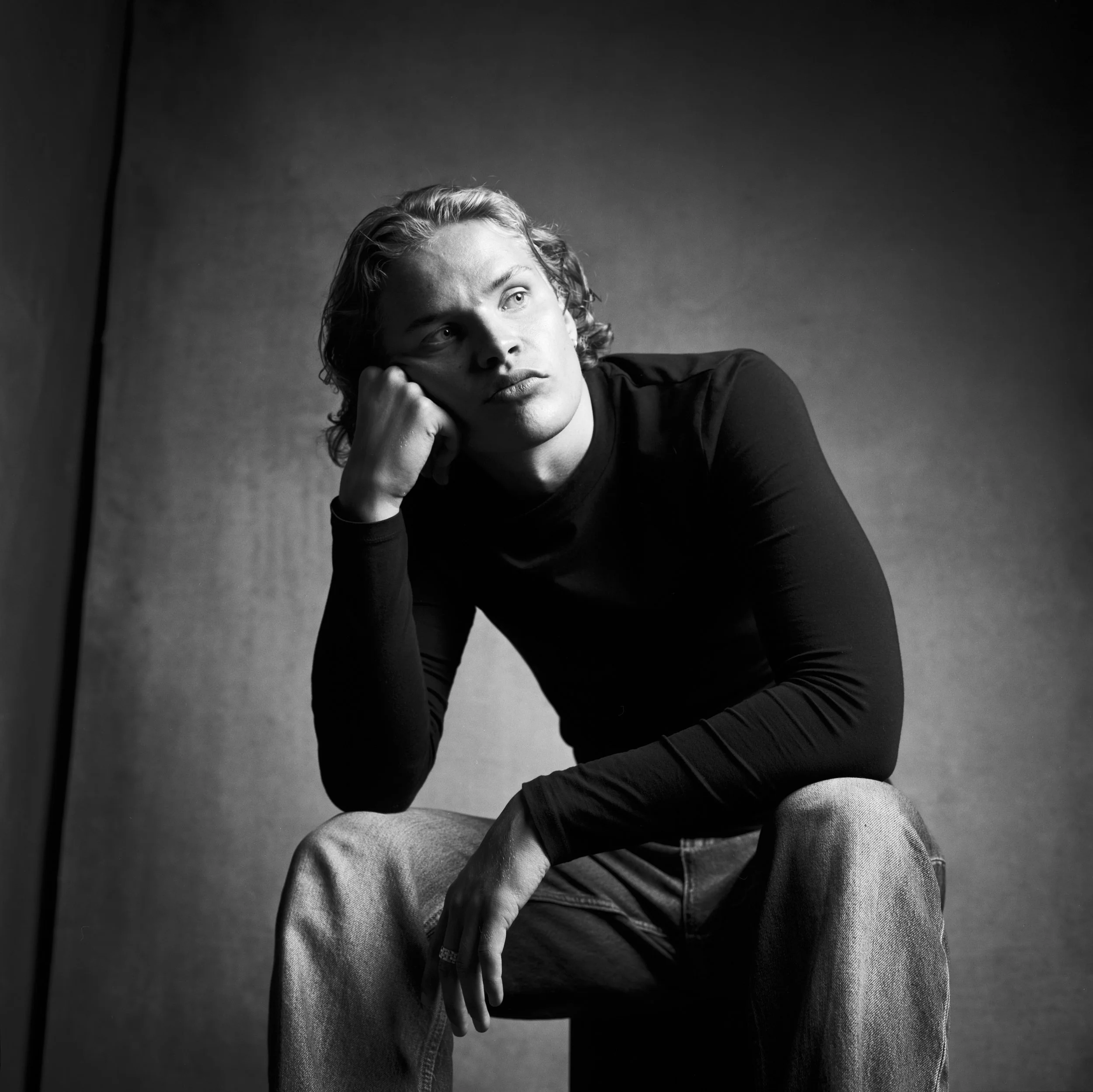 Black and white photo of a young man with curly hair, resting his head on his hand, sitting against a plain background, looking thoughtfully to the side.