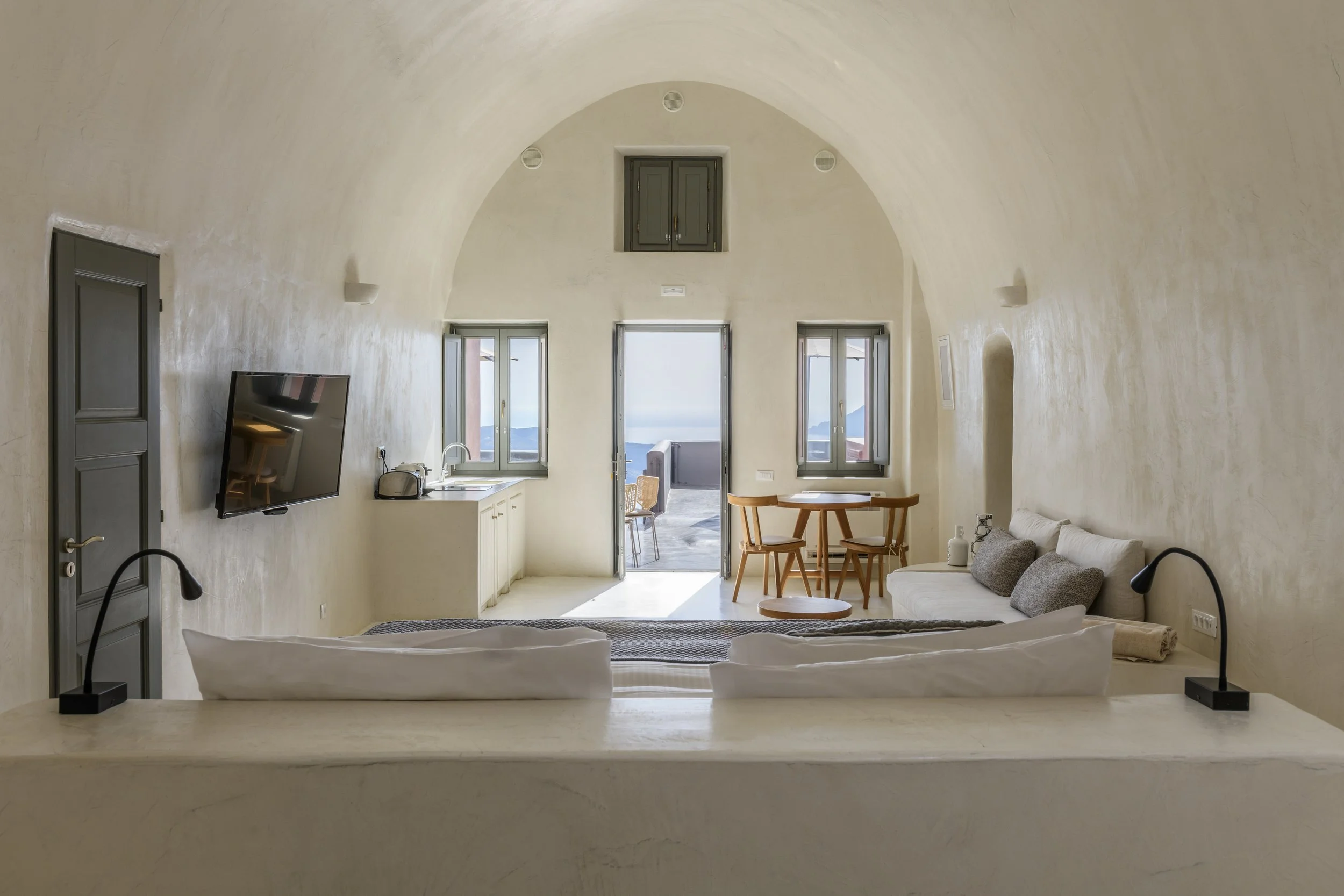 Interior of a room with a curved, white ceiling, three large windows, a small balcony visible outside, a TV mounted on the left wall, a kitchen area with a sink, a table with wooden chairs, a white sofa, and a bed in the foreground.