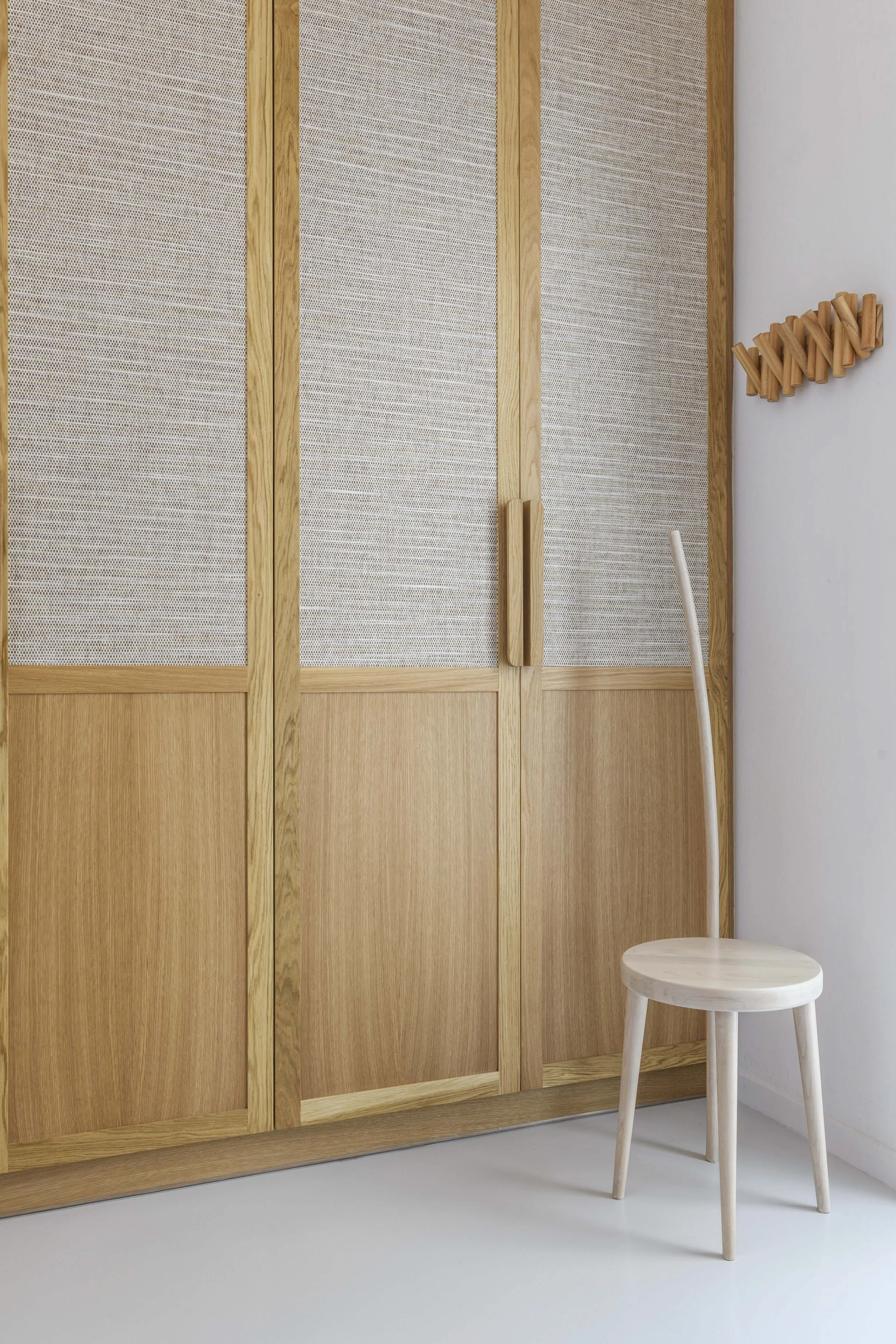 A white wooden chair with three legs and a round seat, positioned next to a light-colored wooden wardrobe with a textured surface and vertical handles, against a plain white wall.