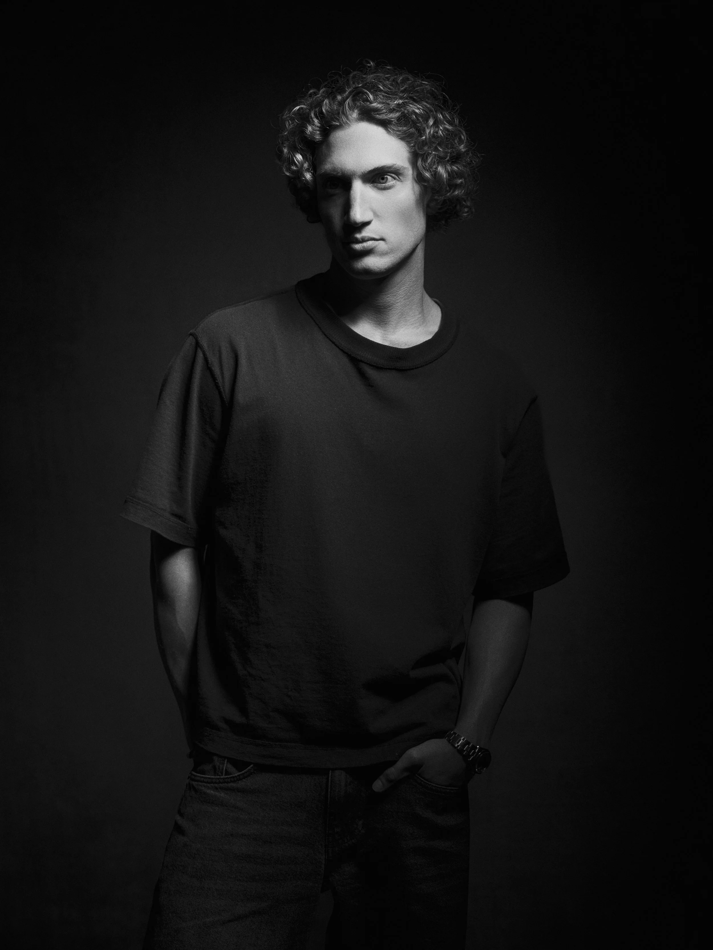 A black-and-white photo of a young man with curly hair, wearing a t-shirt, standing against a dark background. He has a serious expression, with one hand resting near his waist.