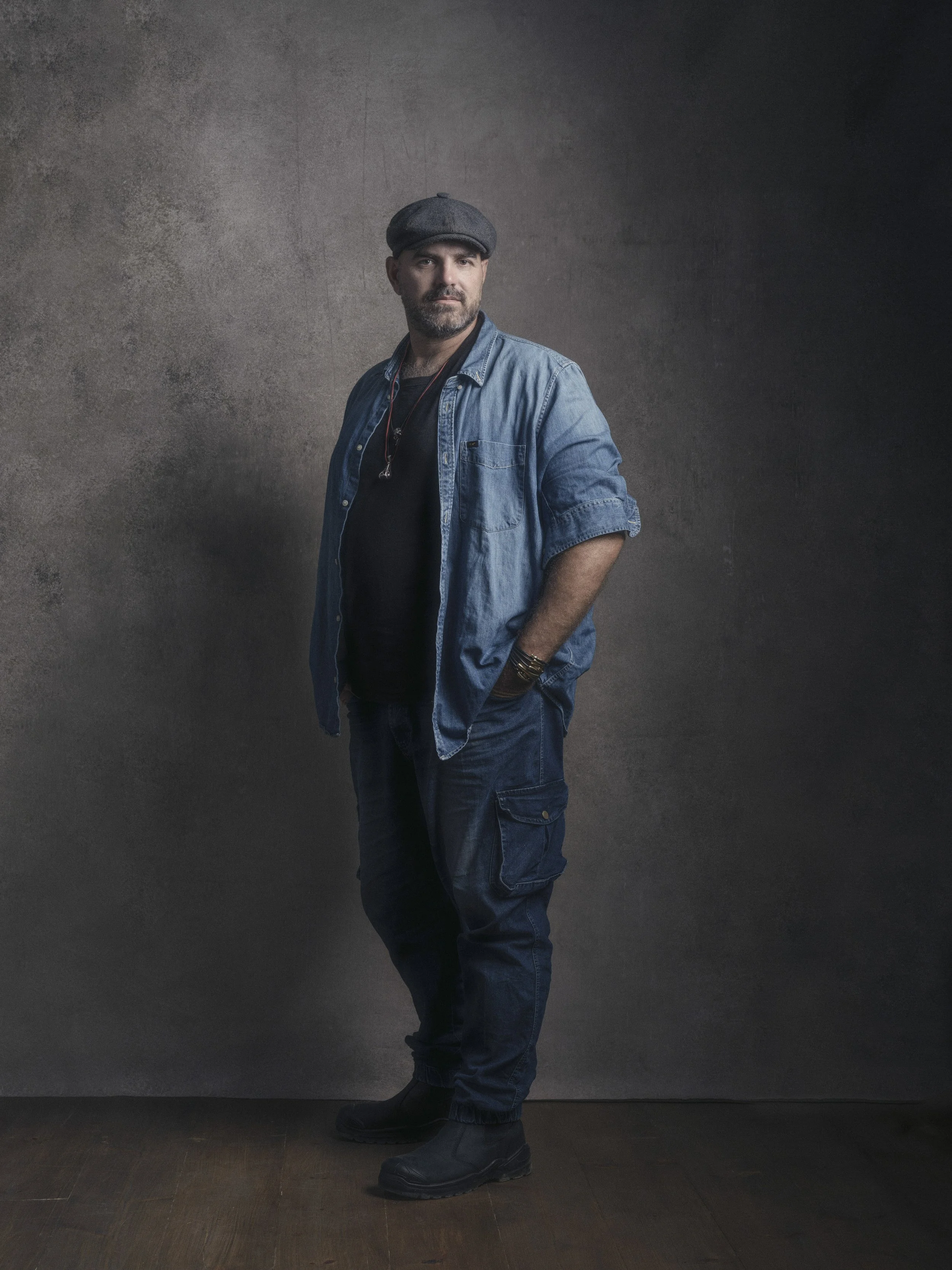 A man wearing a beret, denim jacket, black t-shirt, cargo pants, and boots standing against a textured dark wall.