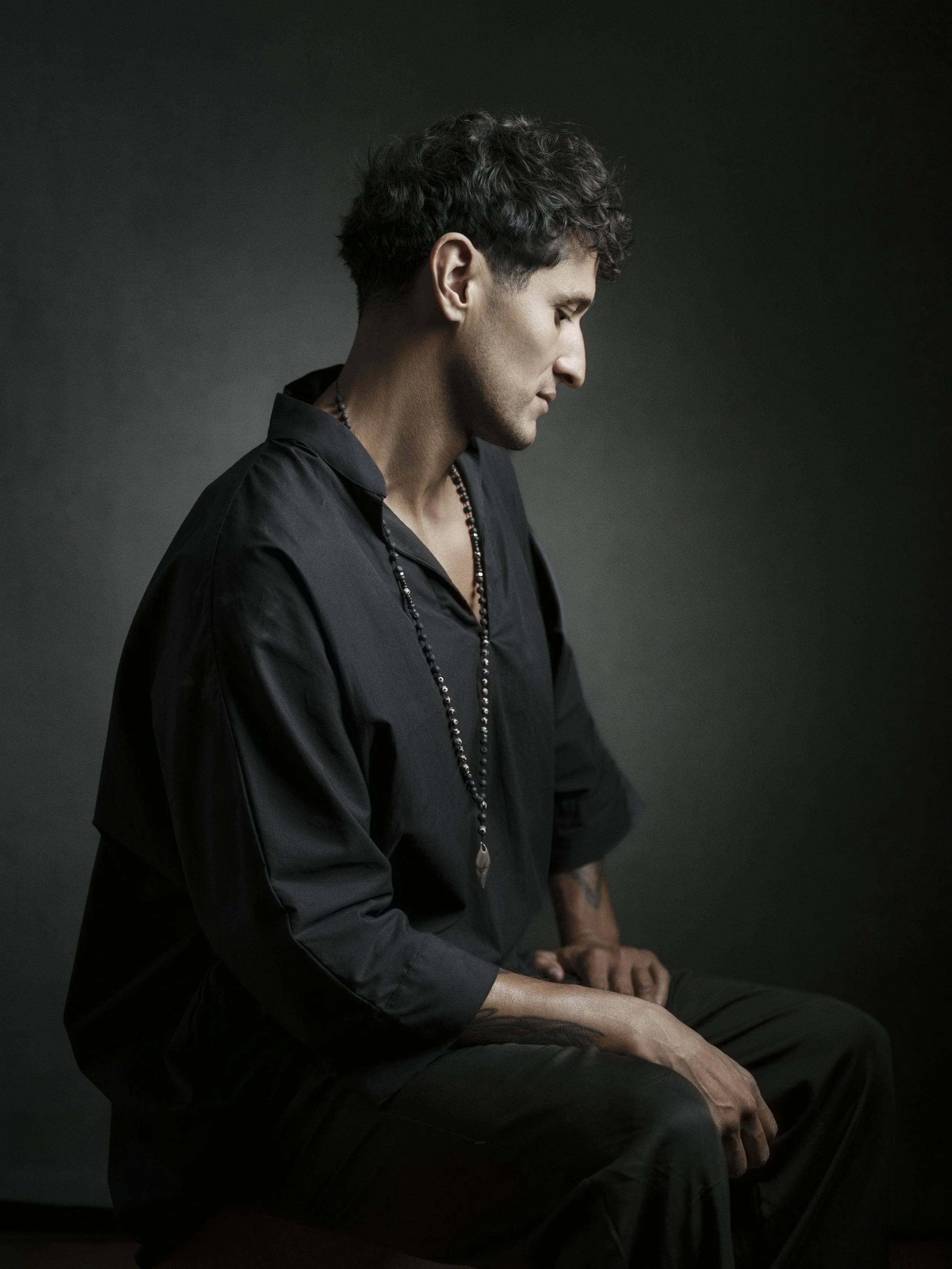 A young man with dark, curly hair and a light complexion, wearing a black shirt and beaded necklaces, sitting against a dark grey background with his eyes closed and head slightly tilted downward.