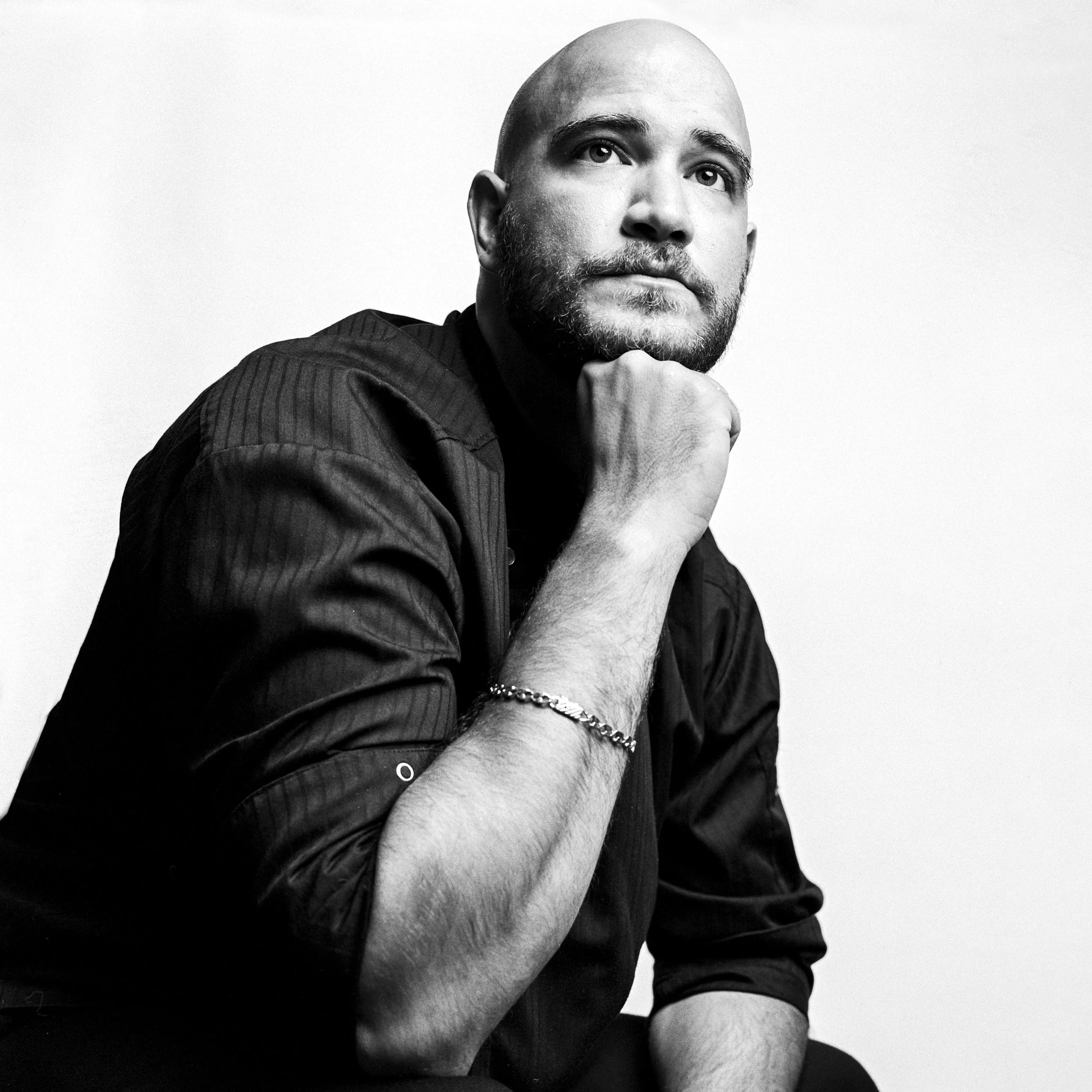 Black-and-white portrait of a man with a shaved head, beard, and mustache, looking upwards thoughtfully with his hand resting on his chin. He wears a dark, textured shirt and a beaded bracelet.