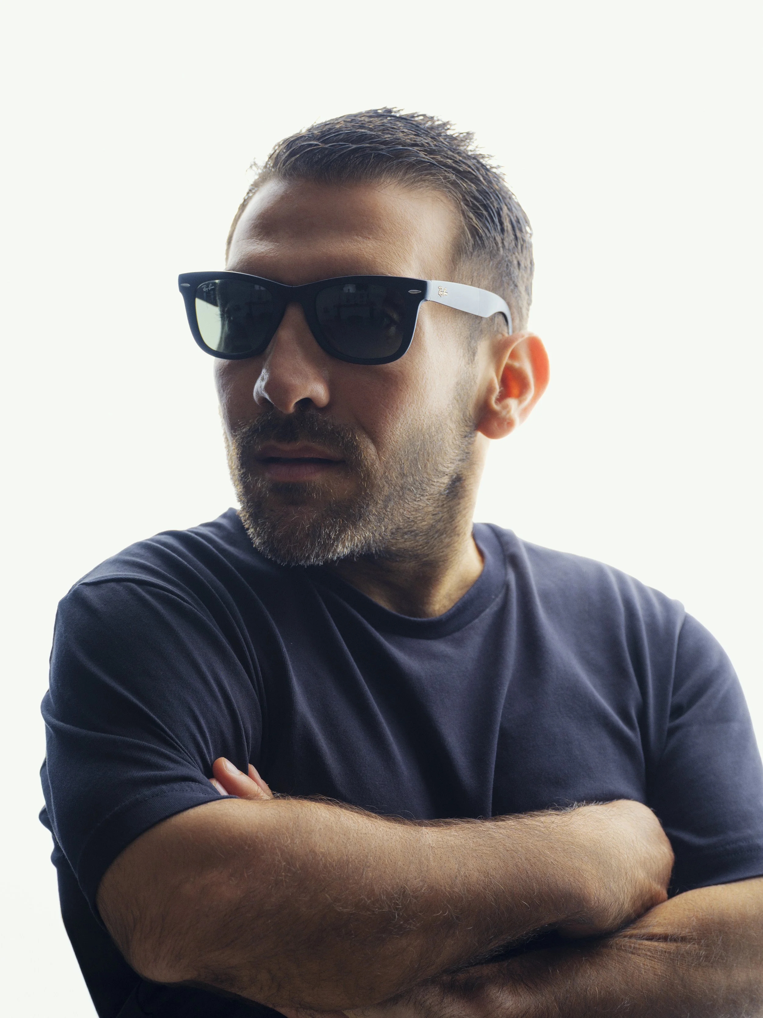 A man with short, styled hair and a beard wearing black sunglasses and a black t-shirt, crossing his arms.