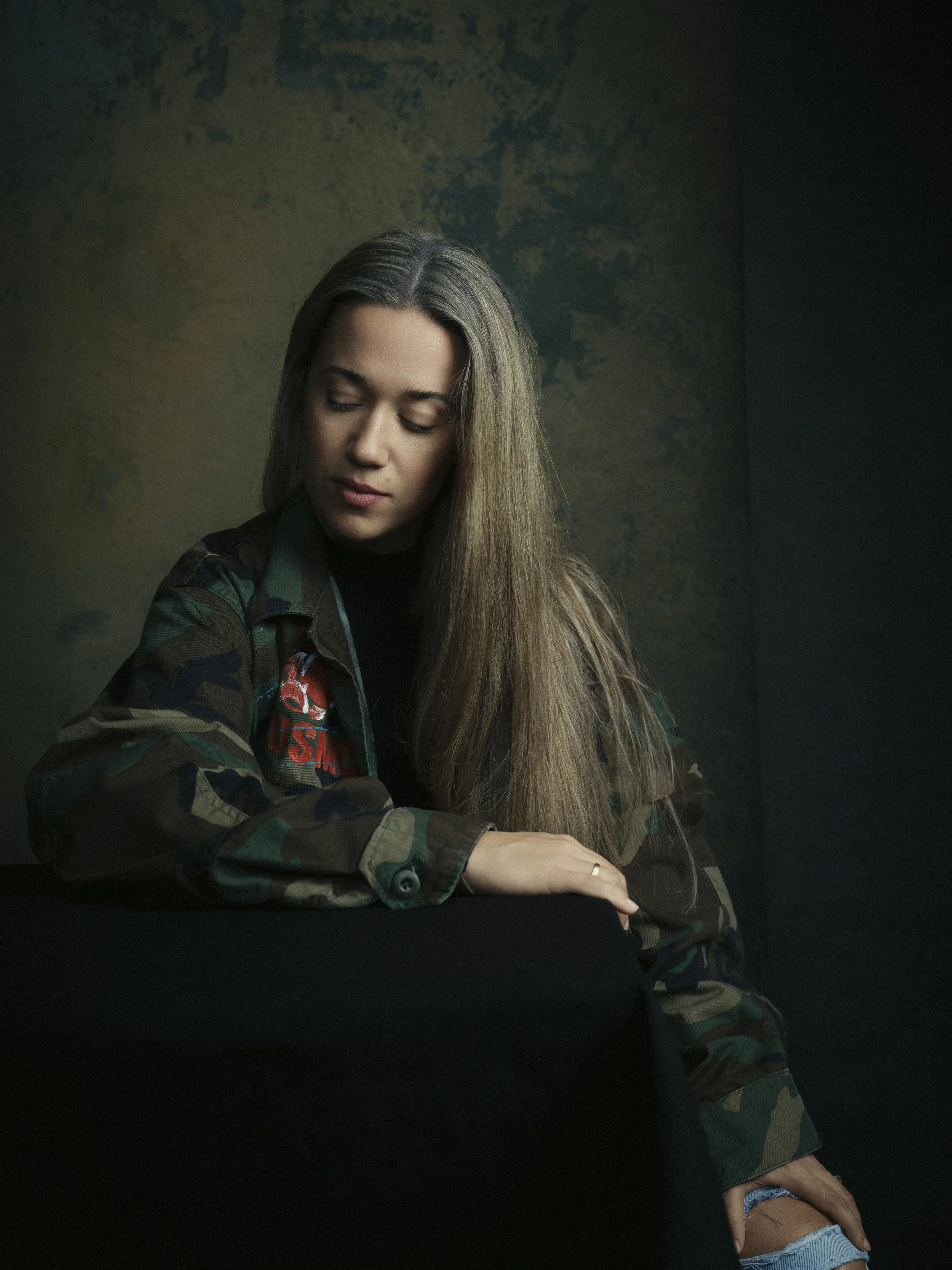 A young woman with long blonde hair in military-style camouflage clothing sits at a black table, looking down with a calm expression. The background is dark with a textured, mottled pattern.