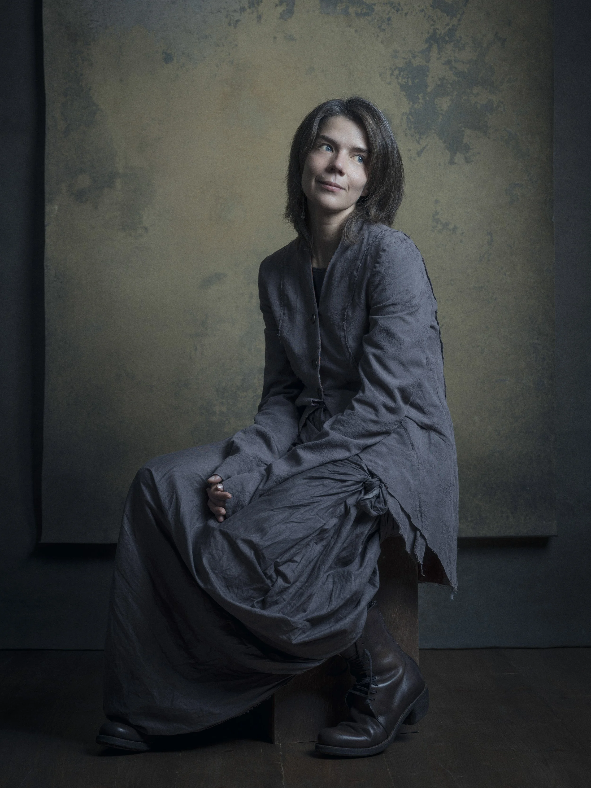A woman with brown hair and blue eyes, wearing a long gray coat and tall black boots, seated against a textured, dark-colored background.