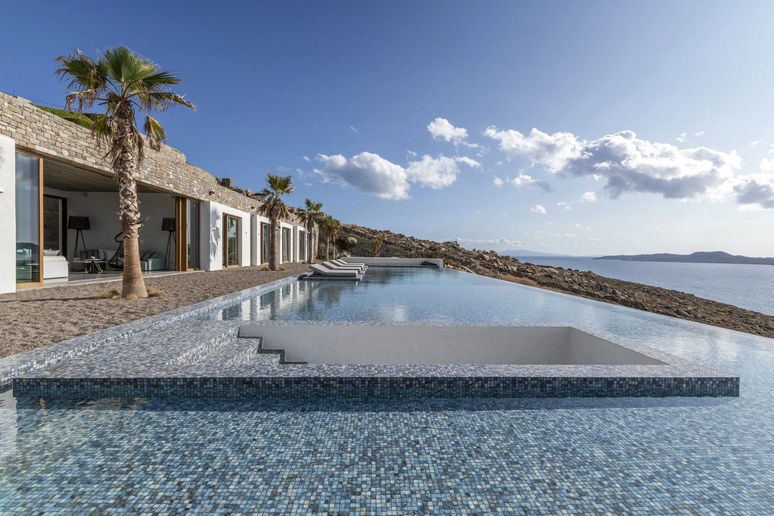 A modern Villa in Mykonos with large glass sliding doors, palm trees, an infinity pool, and a coastal view with islands in the distance under a clear blue sky.