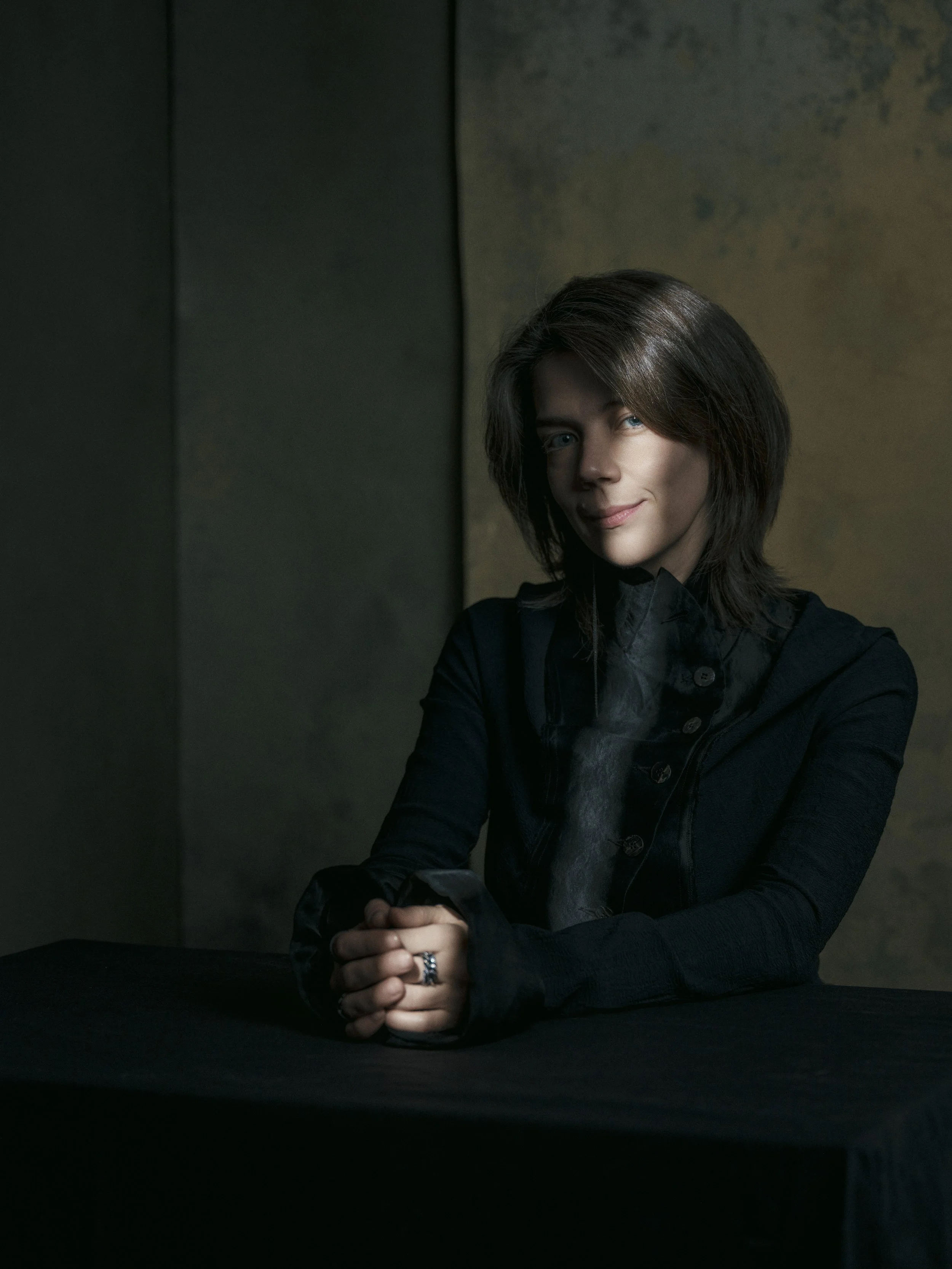 A woman with dark shoulder-length hair and blue eyes is sitting at a dark table with her hands clasped in front of her. She is wearing a black jacket and a dark shirt, and she is smiling softly at the camera. The background is dark with a textured wa