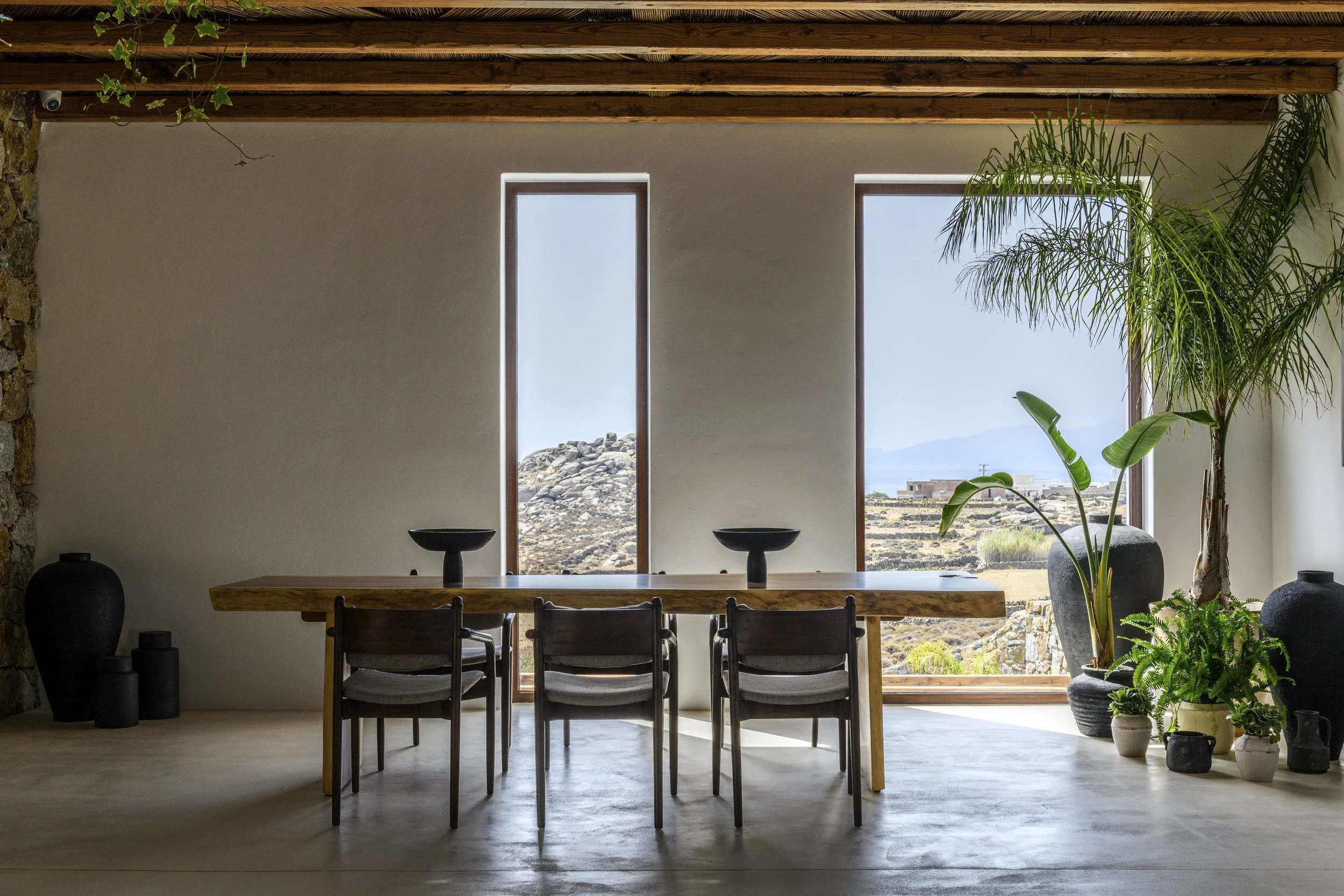 Interior of a modern home with a wooden dining table, six chairs, large windows showing rocky landscape and mountains, and various potted plants.