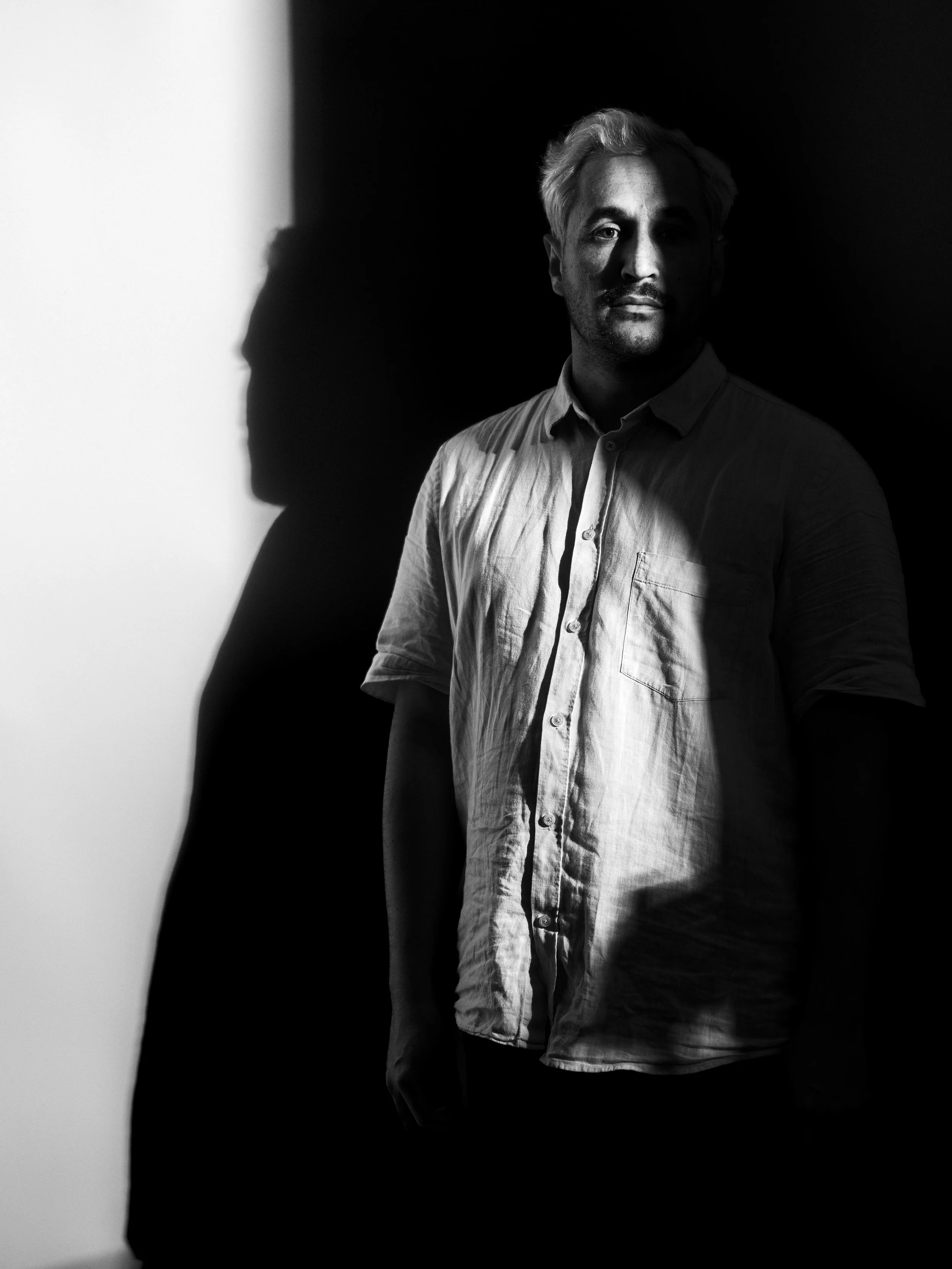 Black and white photograph of a man with light hair, wearing a button-up shirt, standing against a dark background with a white light on one side, casting shadows on his face.
