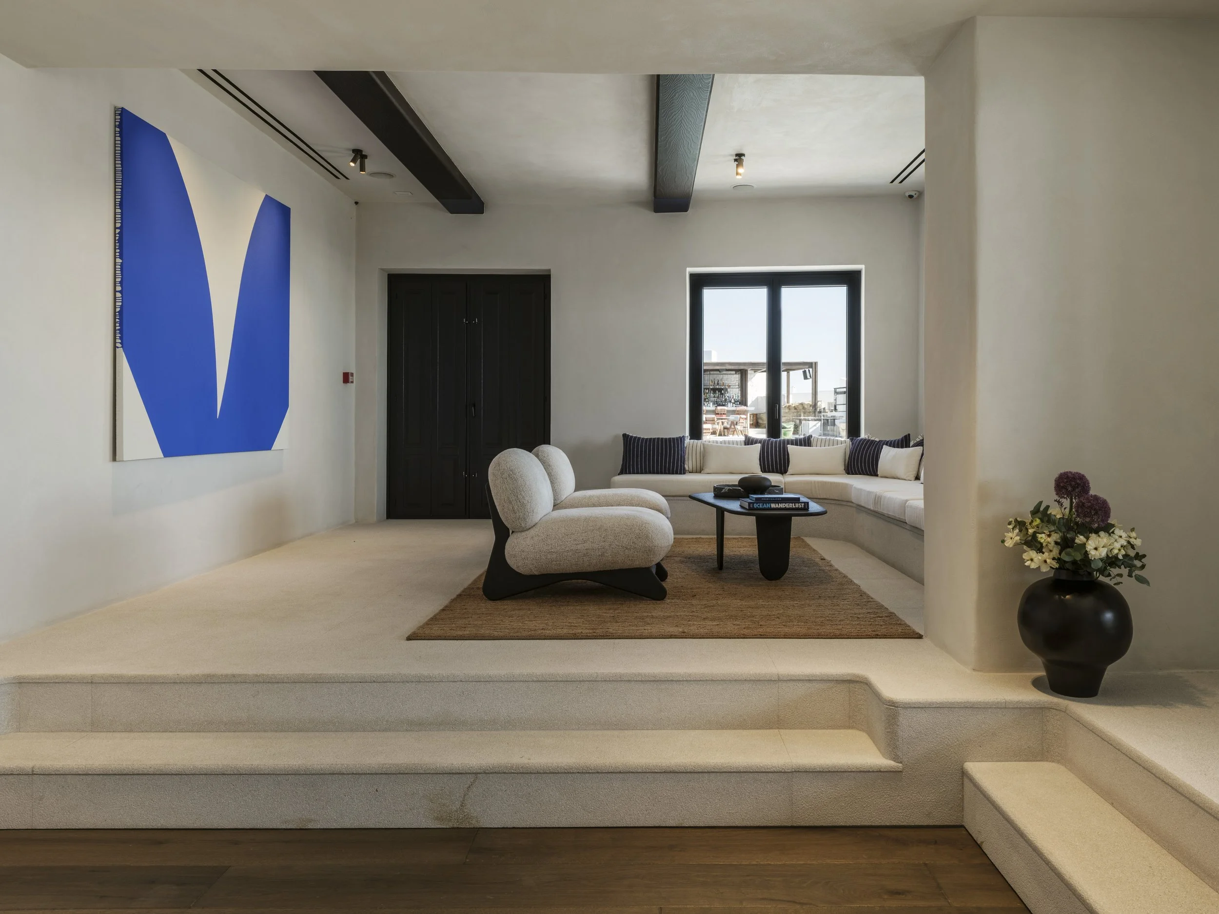 Living room with a white sectional sofa, dark wood window frame, black and white striped pillows, beige armchair, large painting with blue shapes, glass doors leading to an outdoor balcony with table and chairs, potted flowers, hardwood floor.