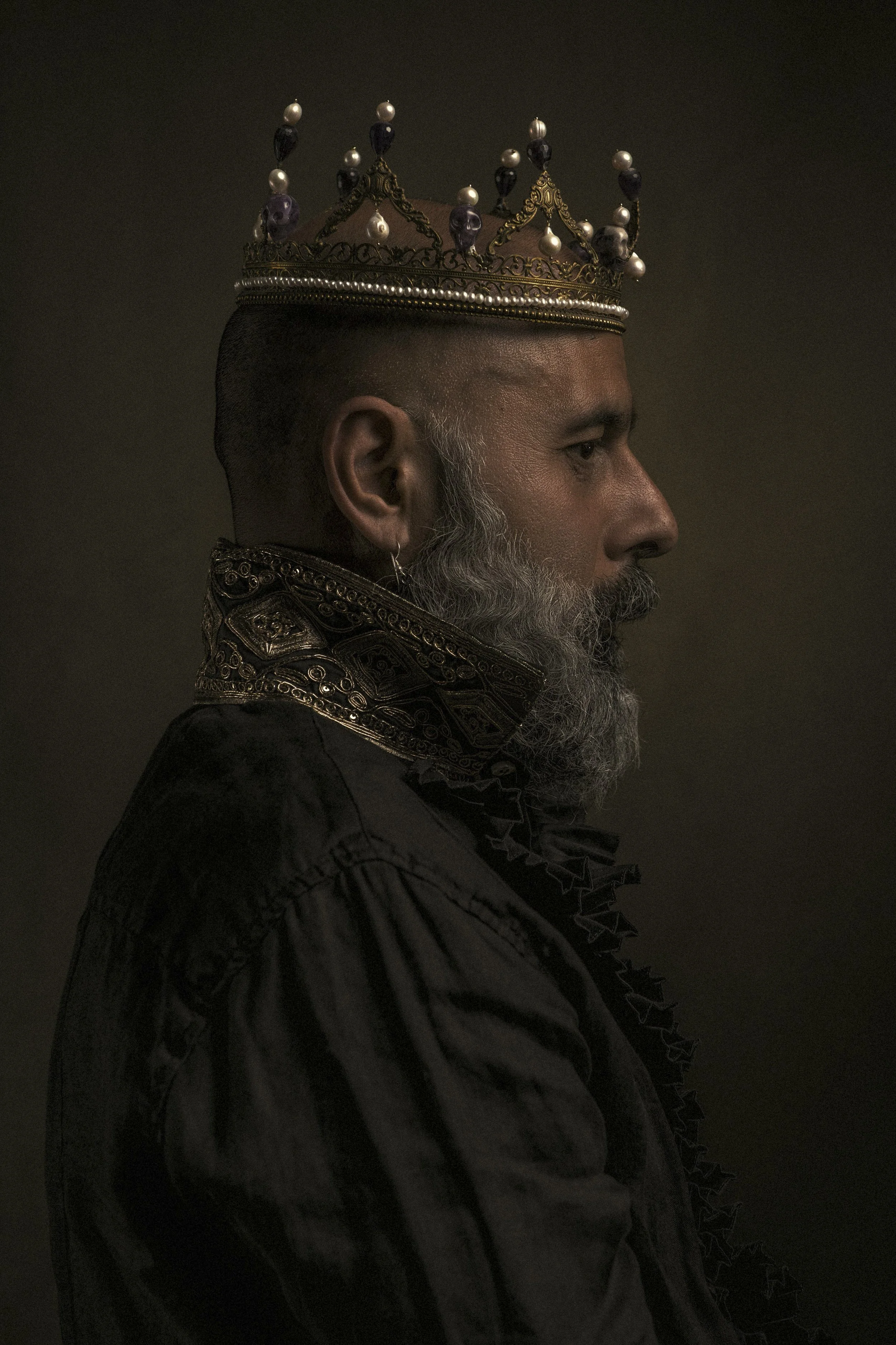 A side profile of a man with dark skin, gray beard, and gray hair, wearing an ornate crown decorated with pearls and purple gemstones, and a dark embroidered jacket against a dark background.