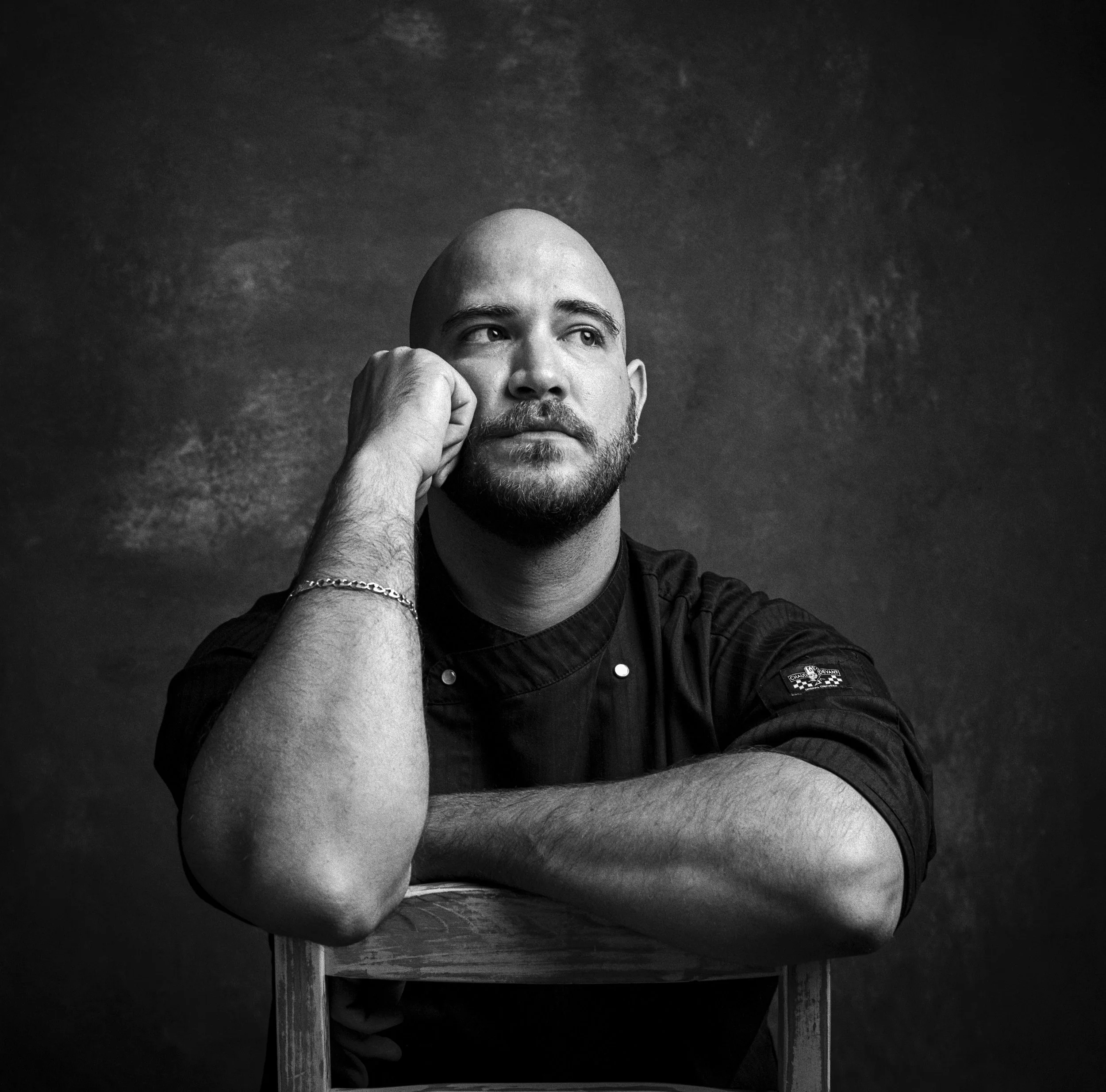 Black and white photo of a man with a shaved head, resting his cheek on his hand, gazing into the distance. He wears a dark shirt with a patch and chain bracelet. The background is dark and textured.