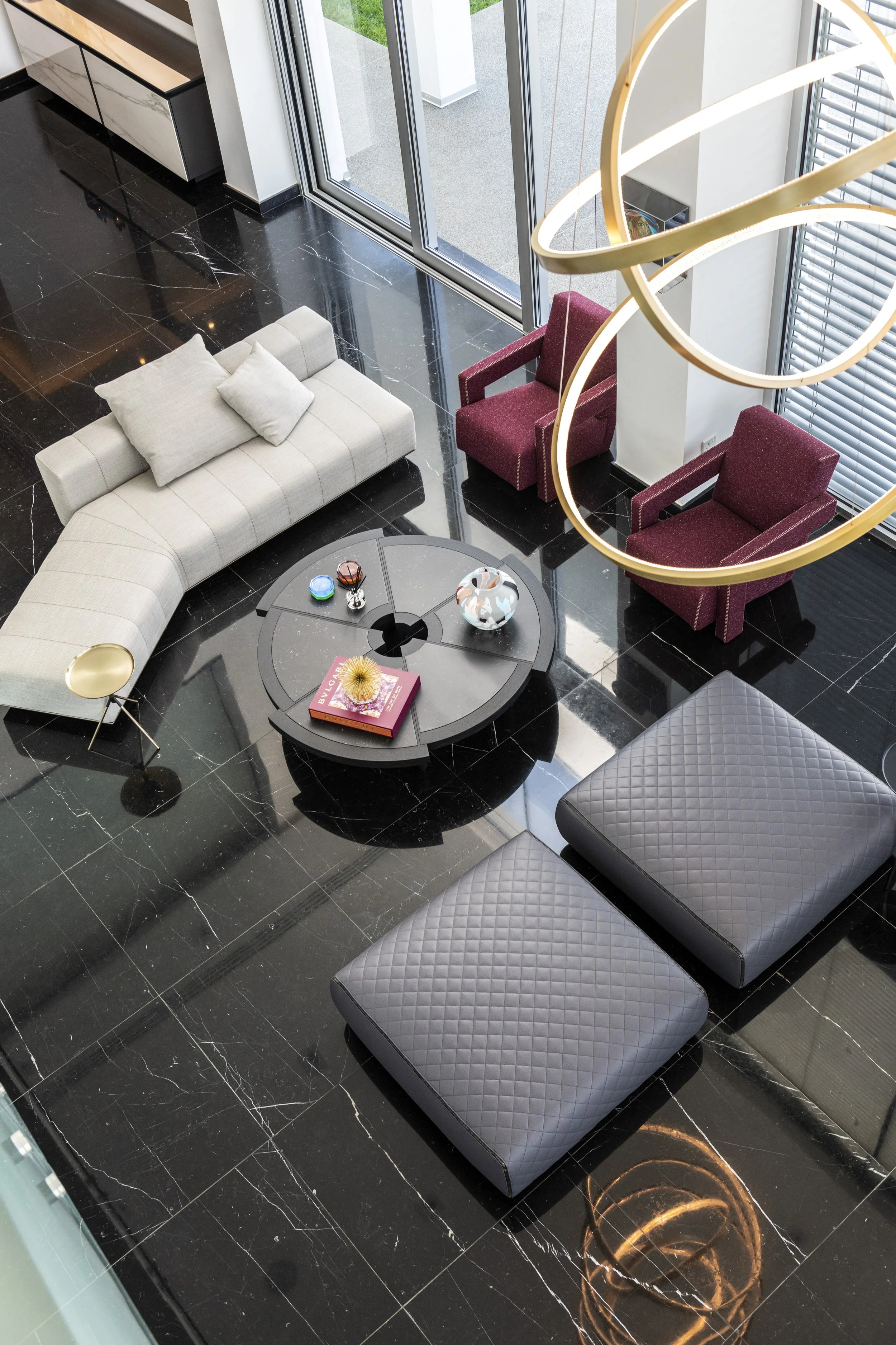 Overhead view of a modern living room corner with black marble floors, a white curved sofa with cushions, two red armchairs, a round black coffee table with decorative objects and books, and a large window with blinds. A gold chandelier with circular