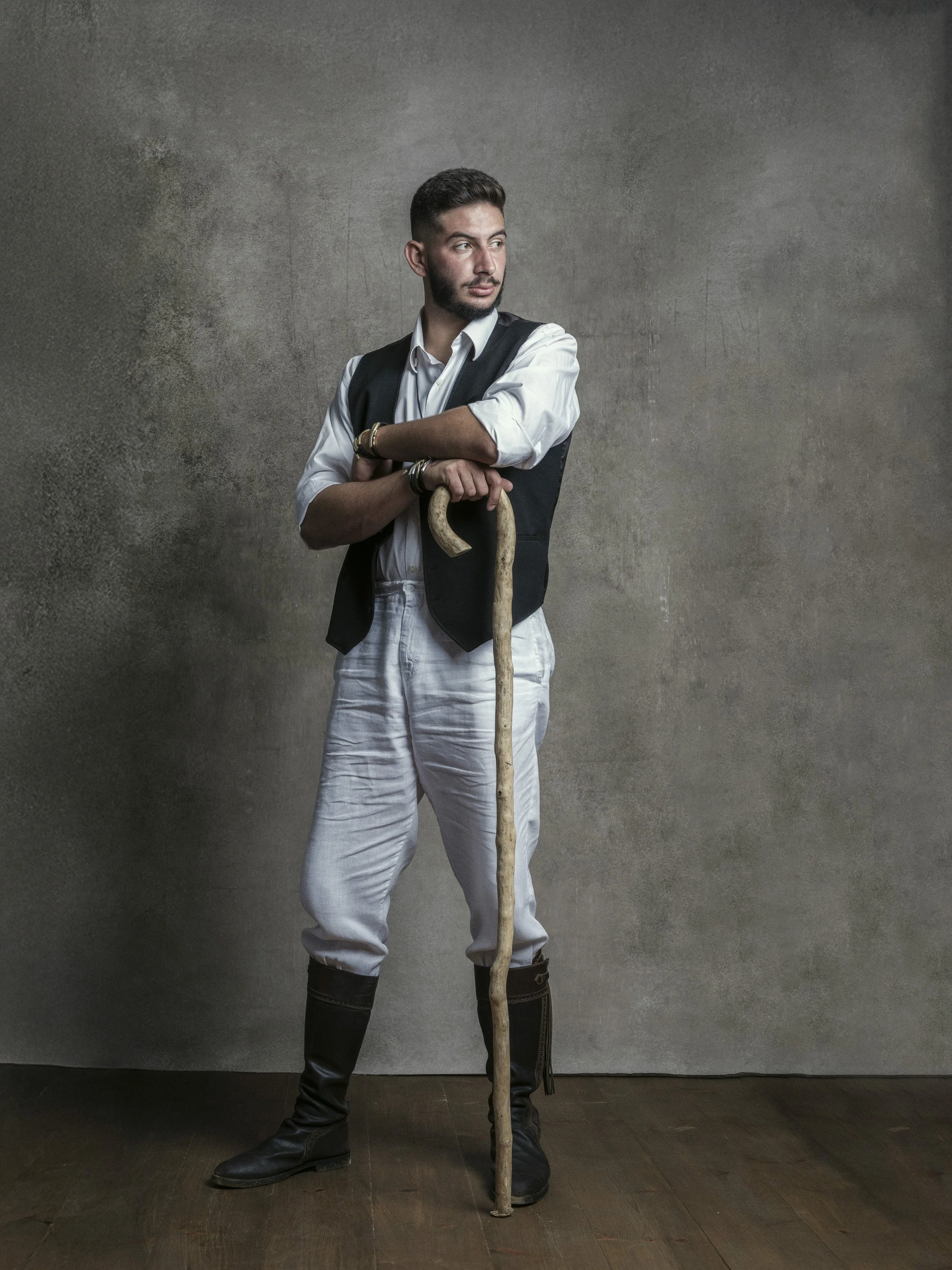 A young man with short dark hair and a beard standing against a textured grey wall, holding a tall wooden staff and looking to the side. He is wearing a white shirt, a black vest, light-colored pants, and black boots.