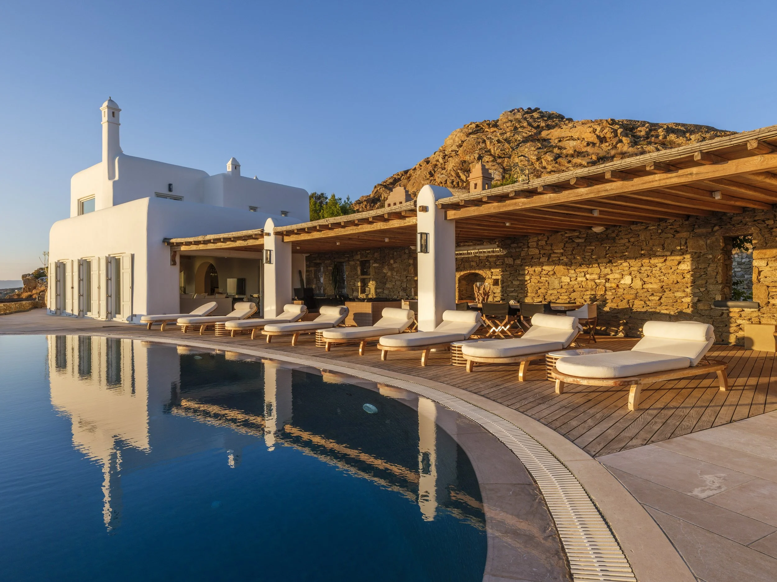 A luxurious outdoor swimming pool with lounge chairs, a white building, wooden pergola, and a rocky hill in the background under a clear blue sky during sunset.