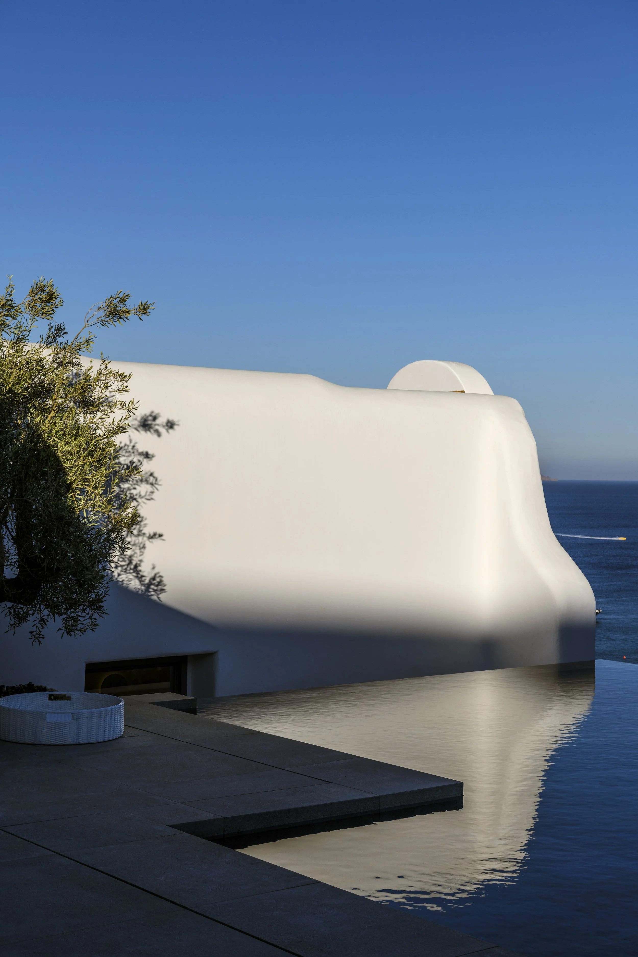 White modern building with curved roof and an outdoor pool reflecting the building, with the ocean and blue sky in the background, and a green tree on the left.