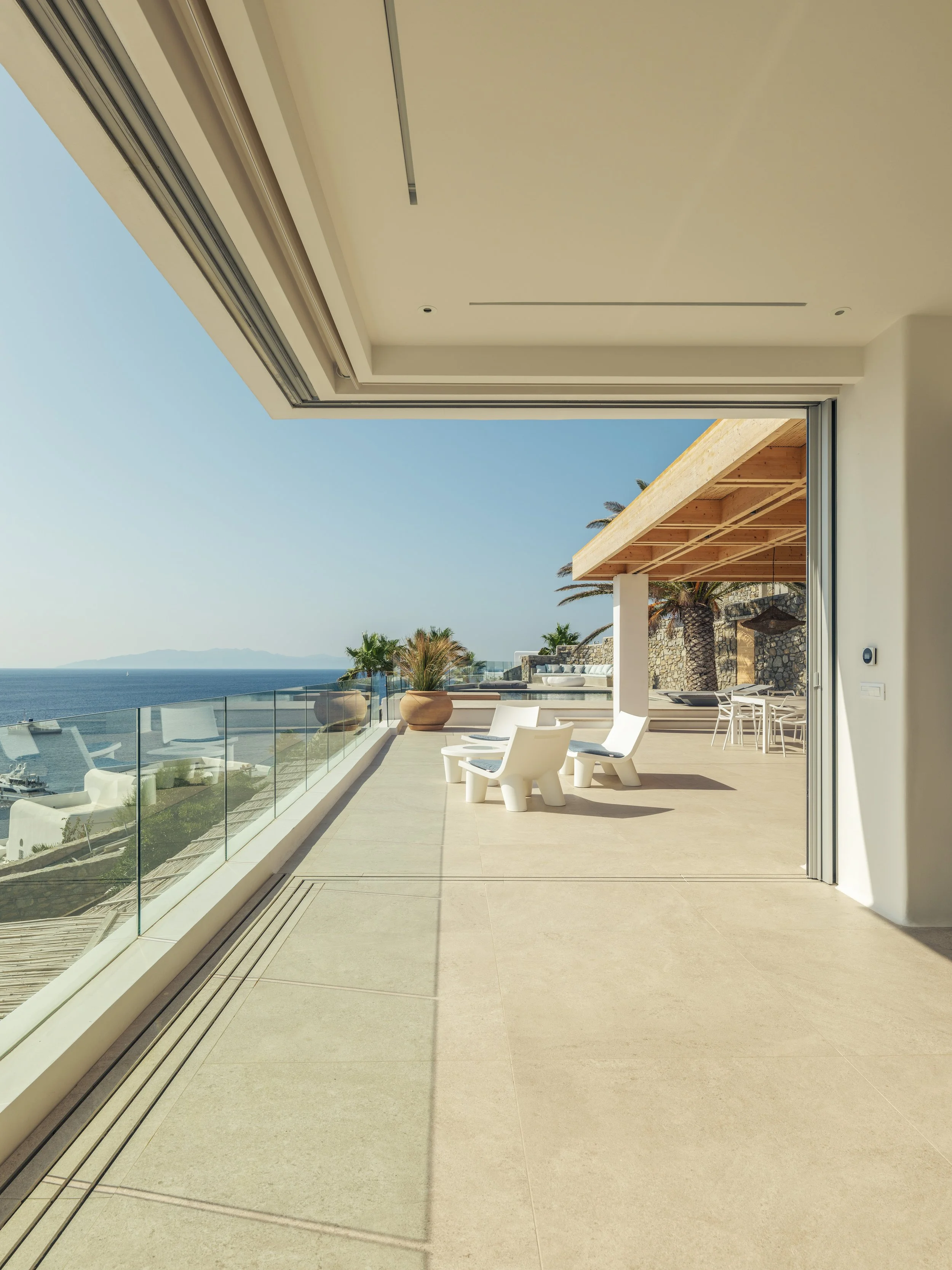 A spacious outdoor balcony overlooking the ocean, with white chairs, potted plants, and a wooden pergola, under clear blue skies.