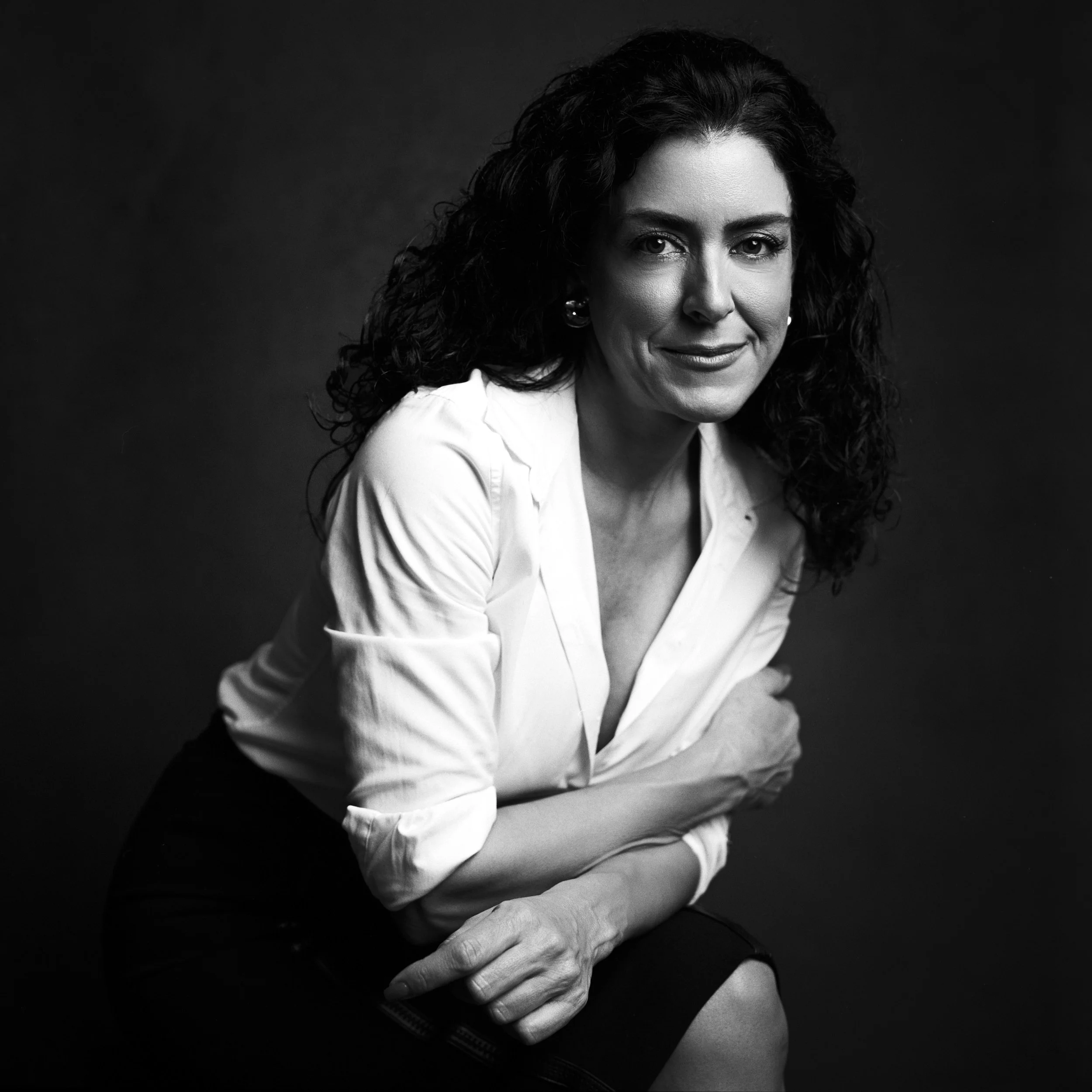 Black and white portrait of a woman with curly hair wearing a white shirt, smiling softly and looking at the camera, against a dark background.