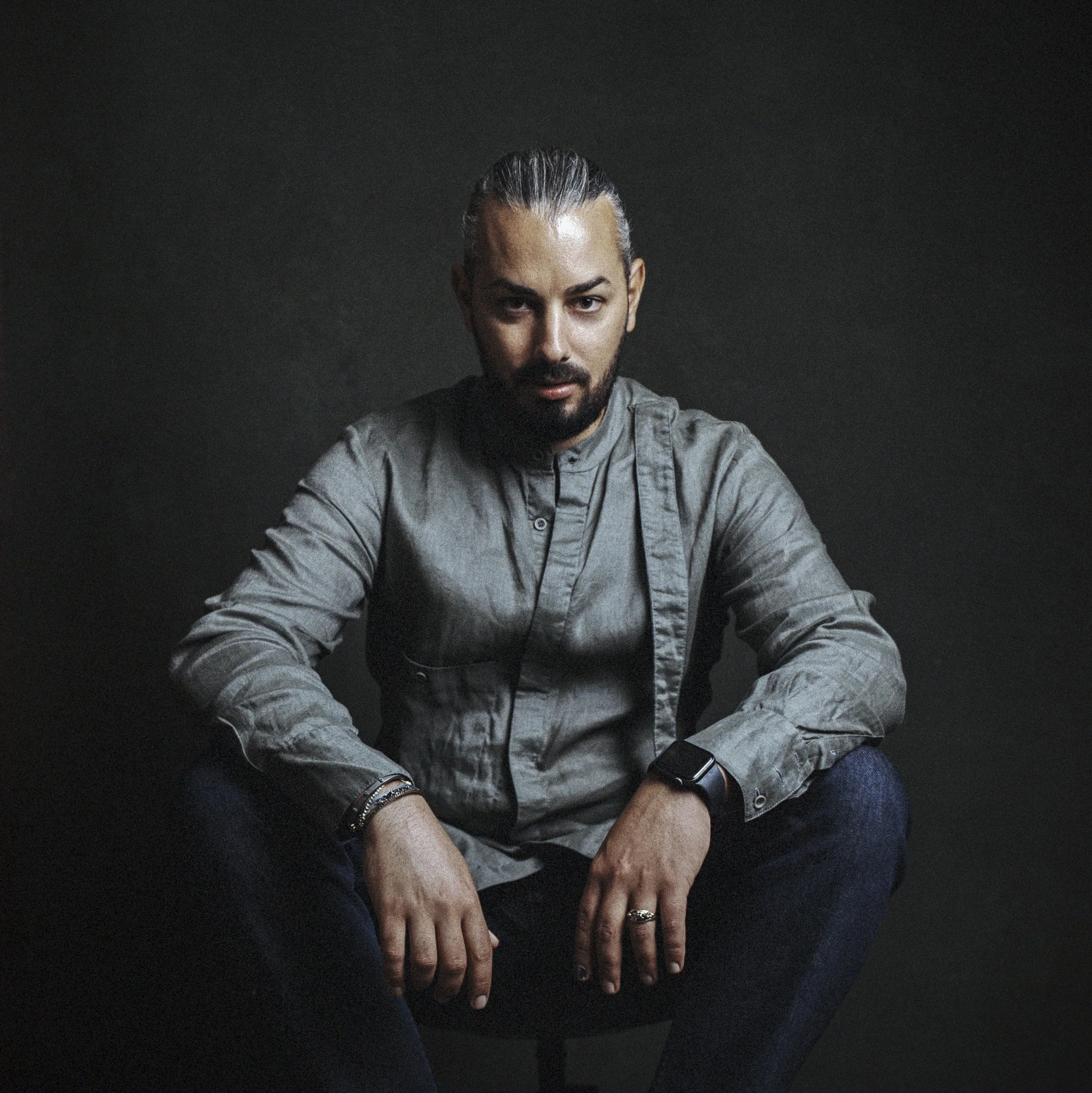 Close-up of a man with a beard and long hair tied back, wearing a gray shirt, sitting with his arms crossed, against a dark background.