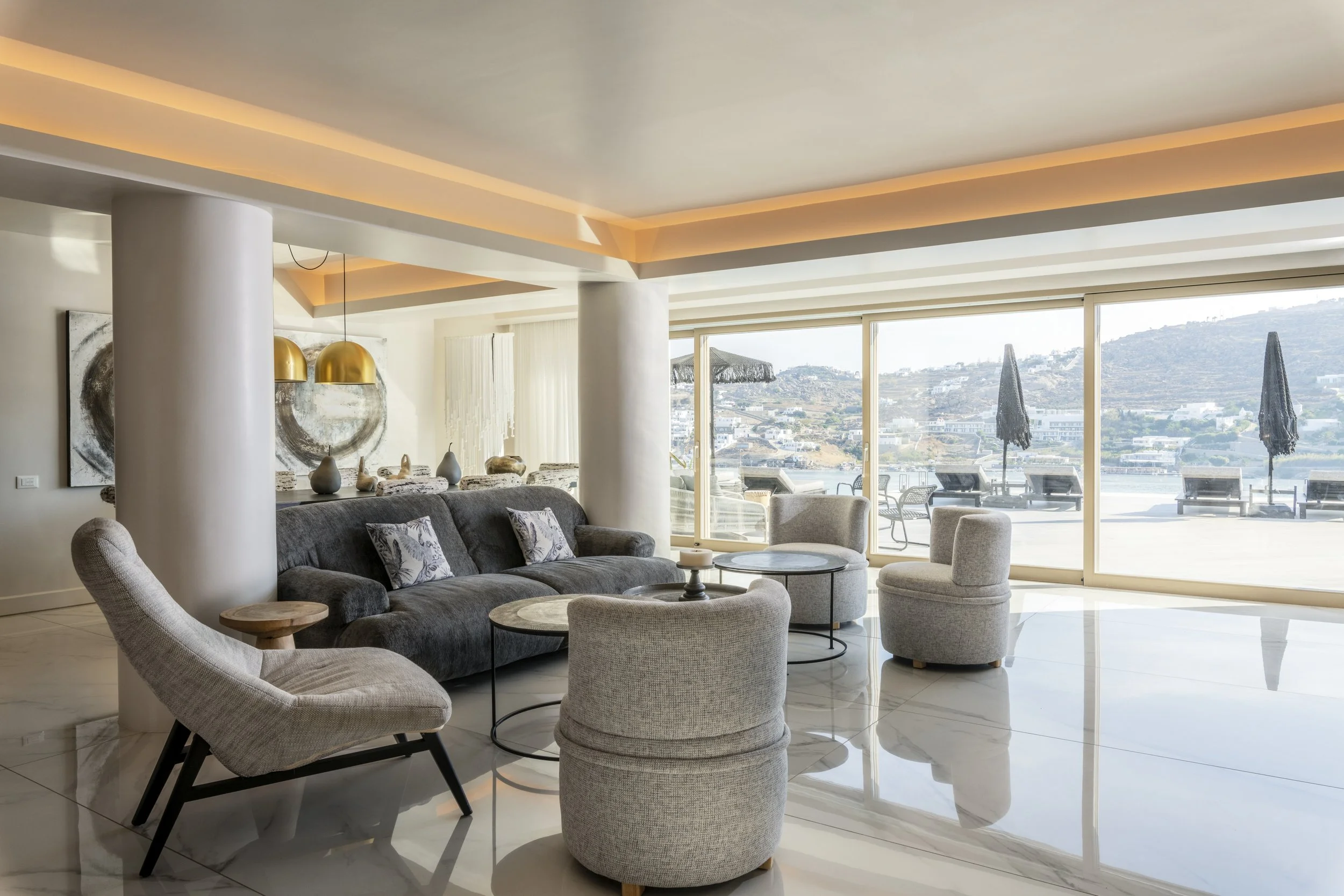 Modern living room with large windows overlooking a hillside landscape, featuring a dark gray sofa, light gray armchairs, white side tables, abstract wall art, and decorative vases, with warm ceiling lighting.