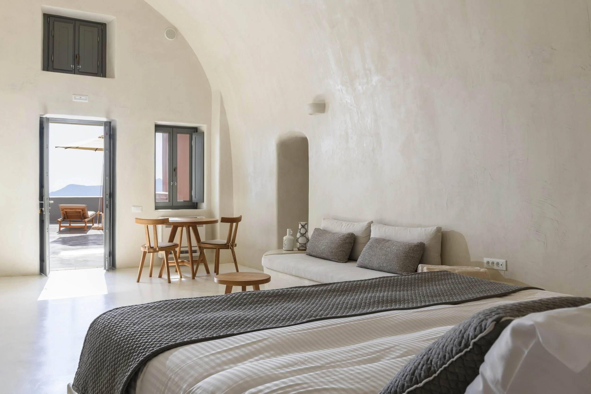 Interior of a minimalist bedroom with a bed, gray and white bedding, and gray pillows. There is a seating area with a white couch and gray pillows, a small wooden table, and a round wooden coffee table. The room has tall windows and a door leading to