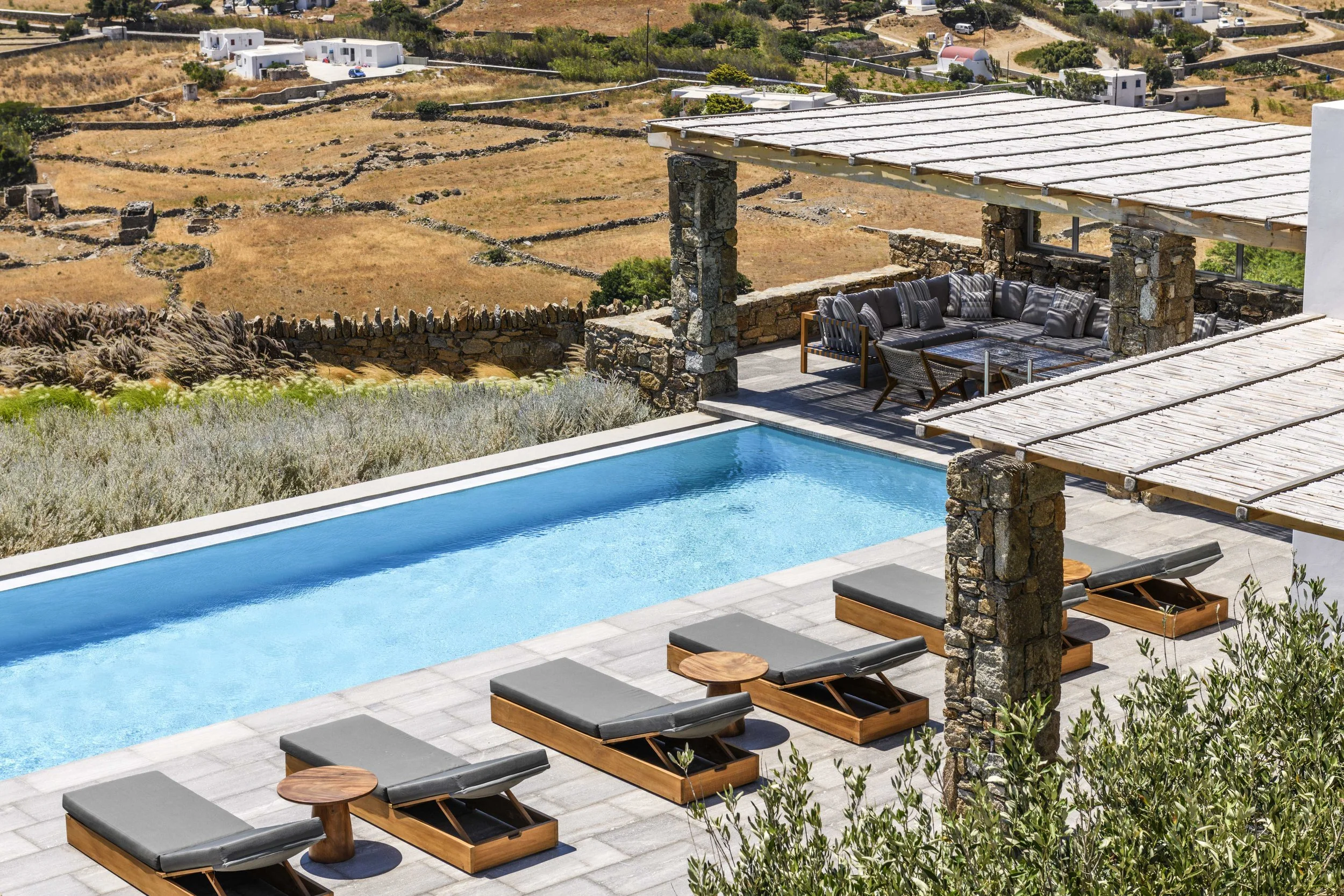 A backyard with a swimming pool, sun loungers, and a shaded seating area with a stone structure. In the background, dry landscape with scattered small white buildings and winding stone walls.