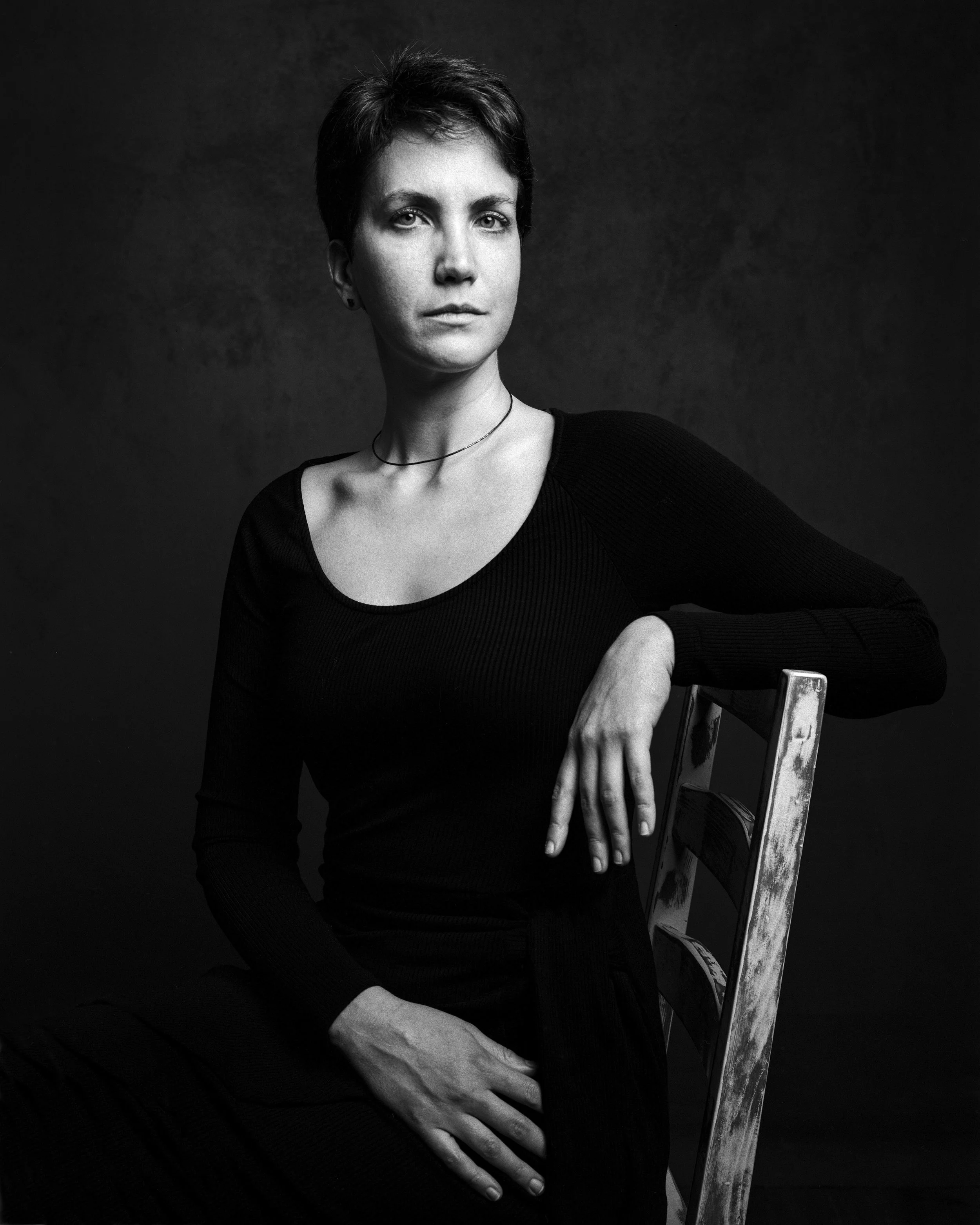 A woman with short dark hair posing with one arm resting on a chair, wearing a black top and a thin necklace, against a dark background in a black-and-white photograph.