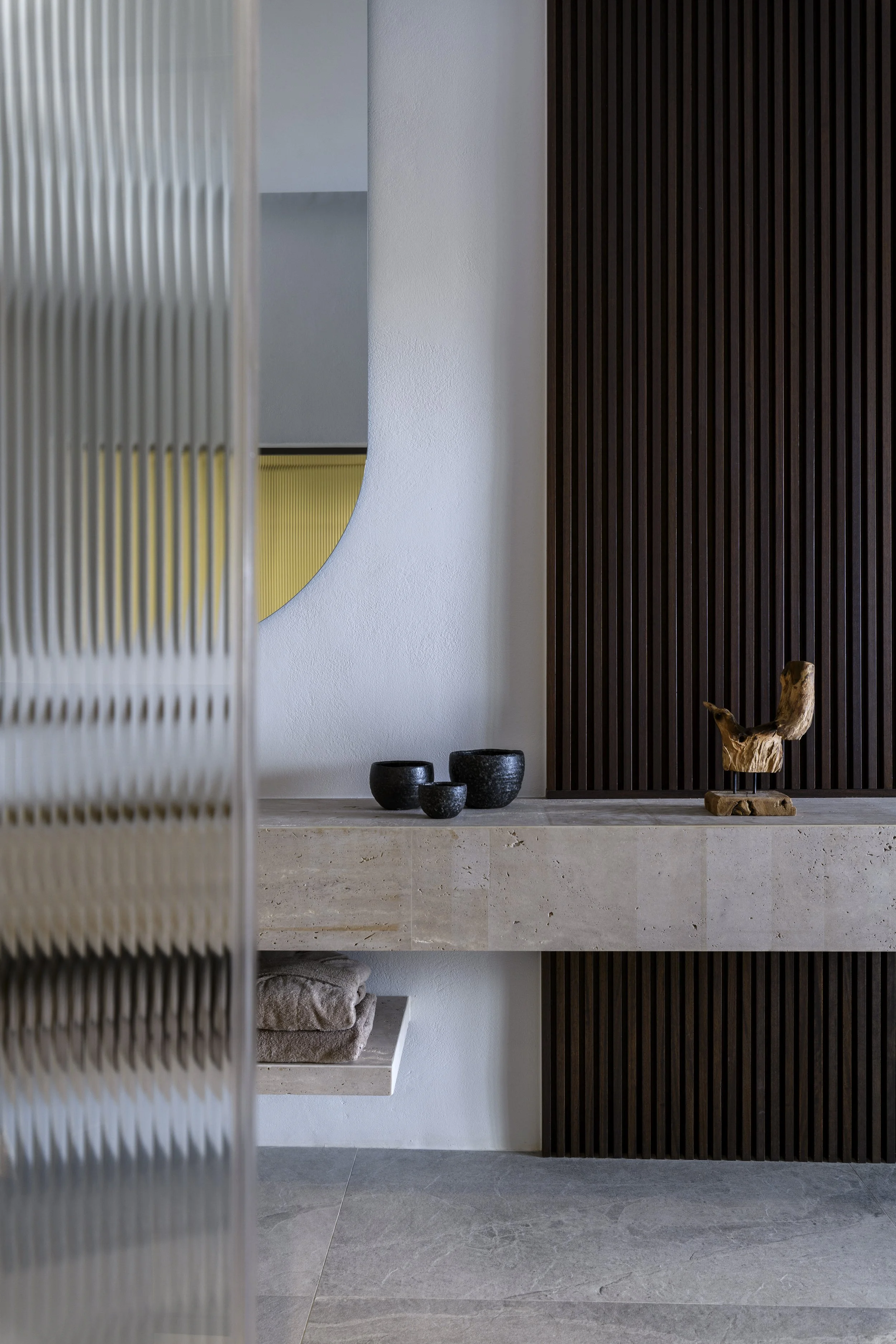 Interior with a minimalist design, featuring a cream-colored stone shelf with neatly folded towels, black ceramic pots, decorative wooden sculpture, a round wall mirror, a dark brown wooden slatted wall, and a white textured wall, with a light gray t