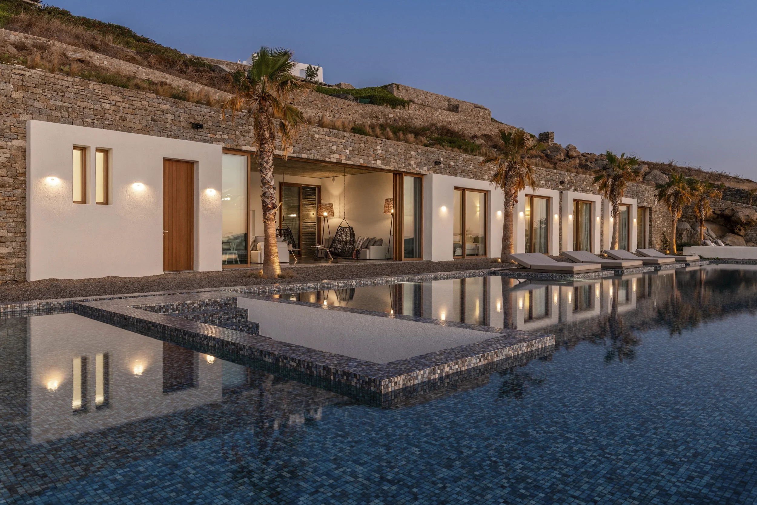 Modern house with large glass windows and doors, palm trees, outdoor seating, and a swimming pool reflecting the house, hillside, and sky at dusk.