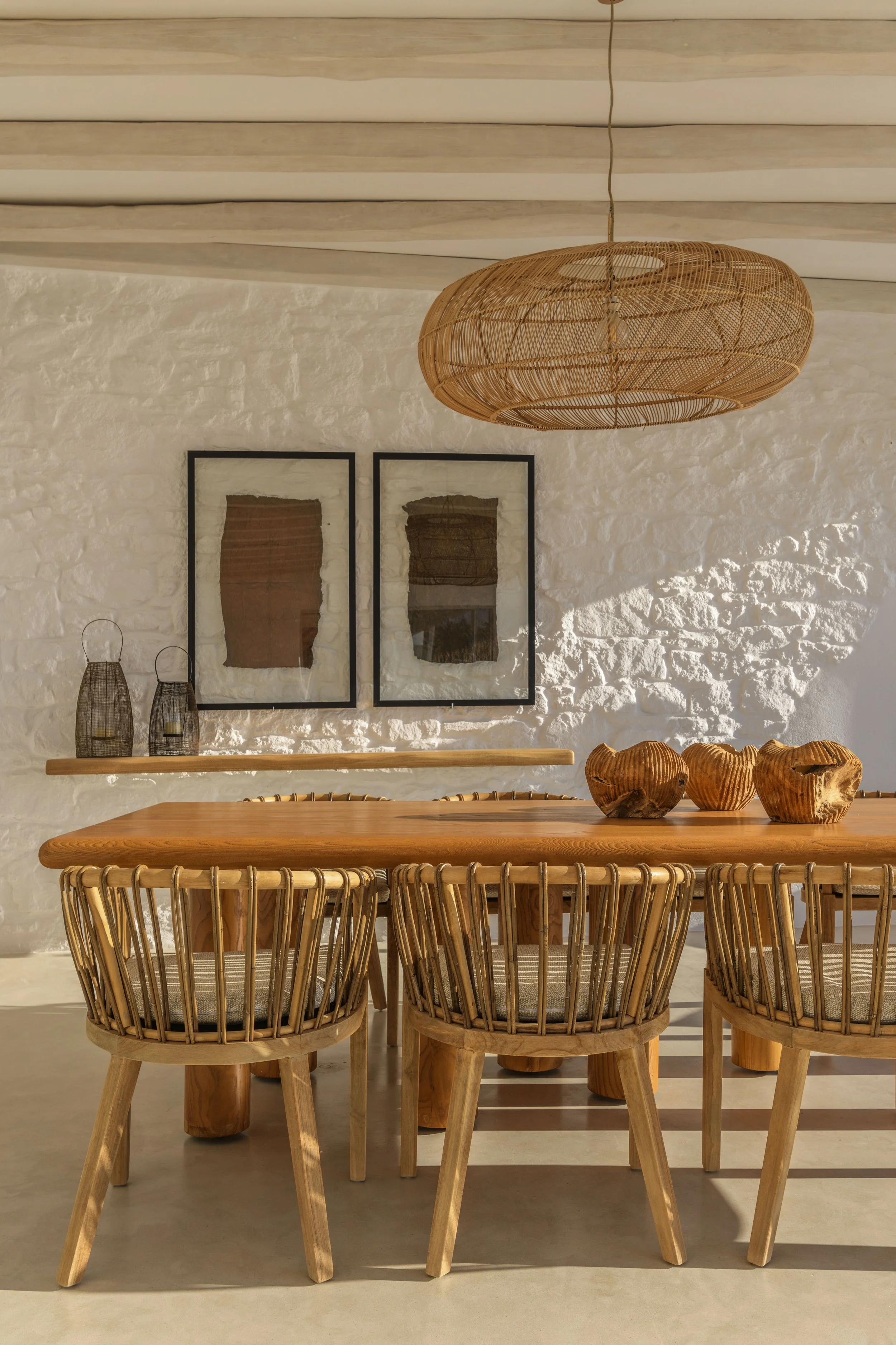 A dining room with a wooden table and six woven wooden chairs. The room has a white textured brick wall with two framed abstract art pieces. Above, a round rattan pendant light hangs from the ceiling.