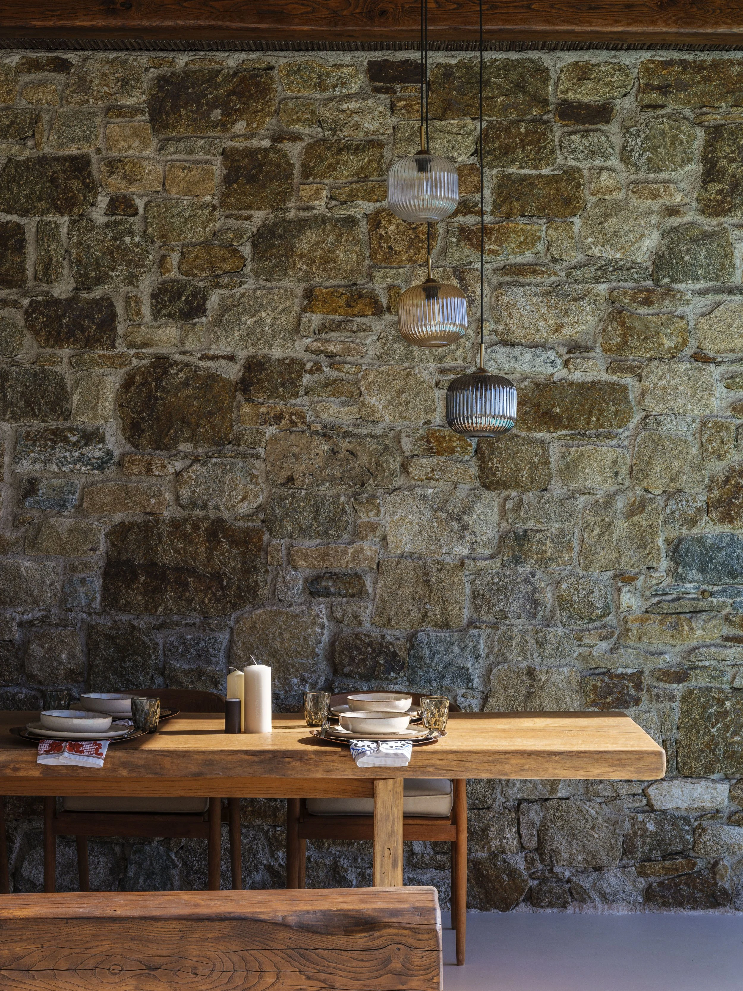 A dining table set with white plates, glasses, and candles against a stone wall with hanging pendant lights.