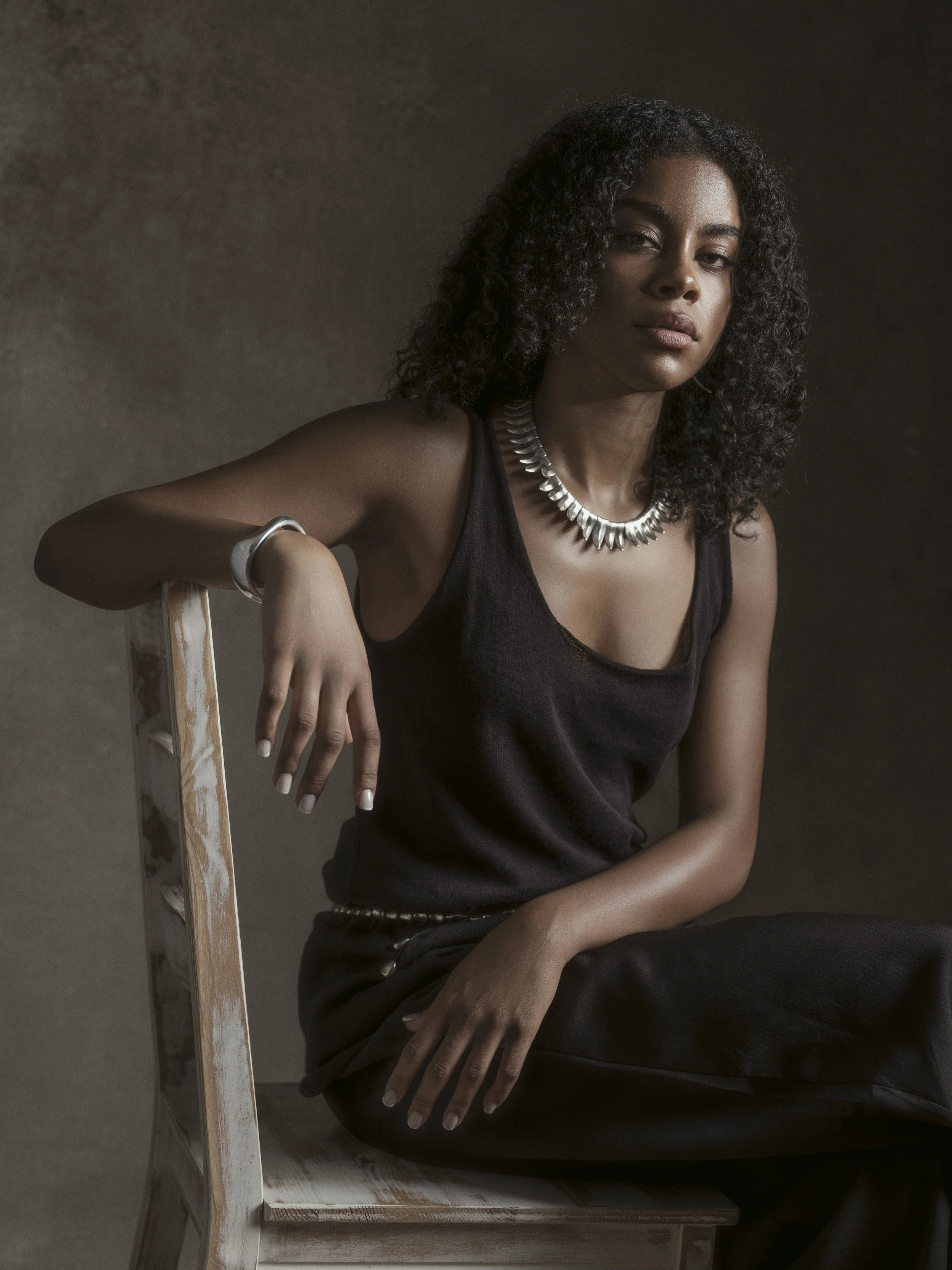 A young woman with dark curly hair and medium skin tone sitting on a distressed white wooden chair against a dark background. She is wearing a black sleeveless top, a silver statement necklace, and a silver bracelet on her right wrist.