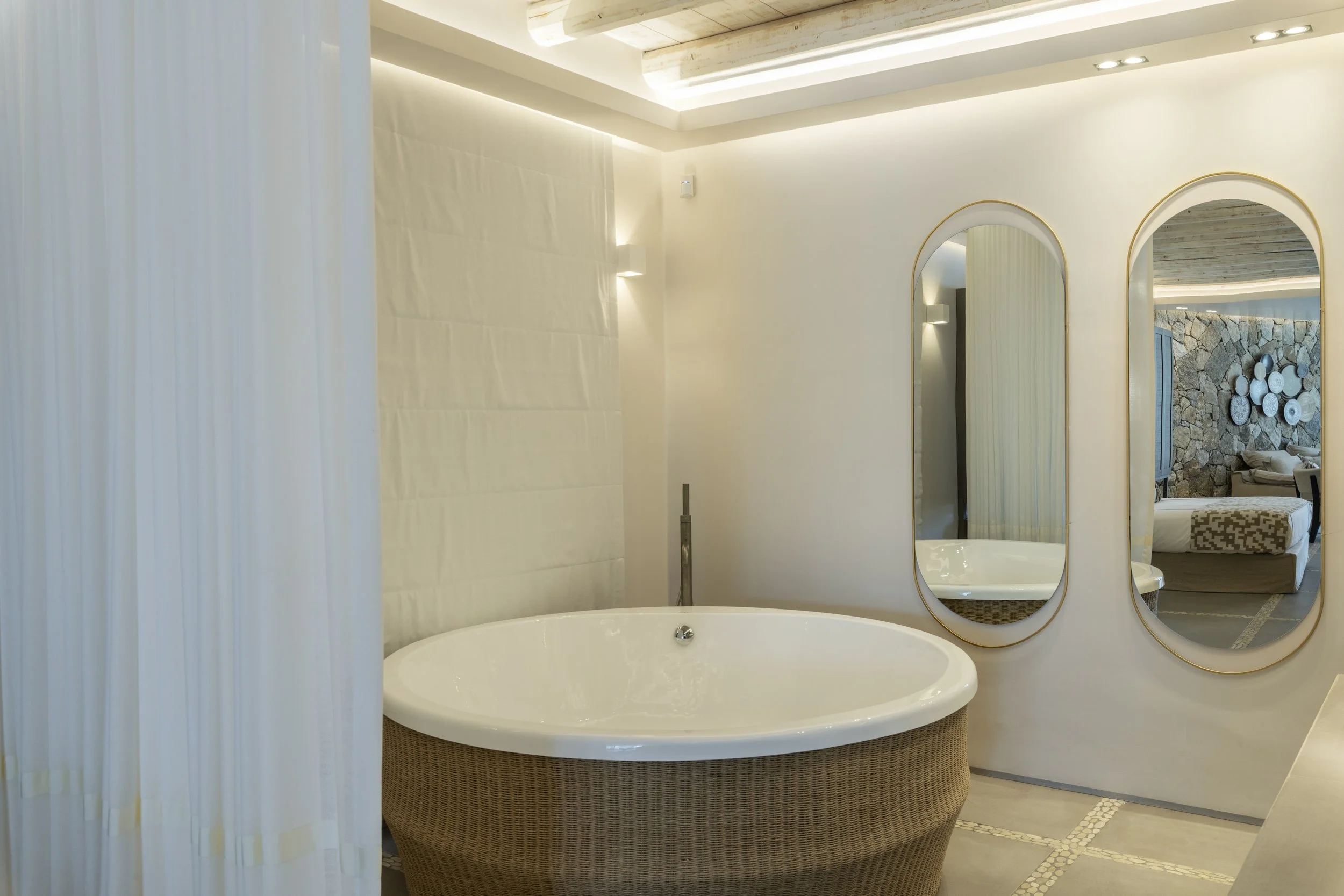 A bathroom with a large round bathtub in front of a wall with two large oval mirrors and a stone accent wall visible through the reflection.