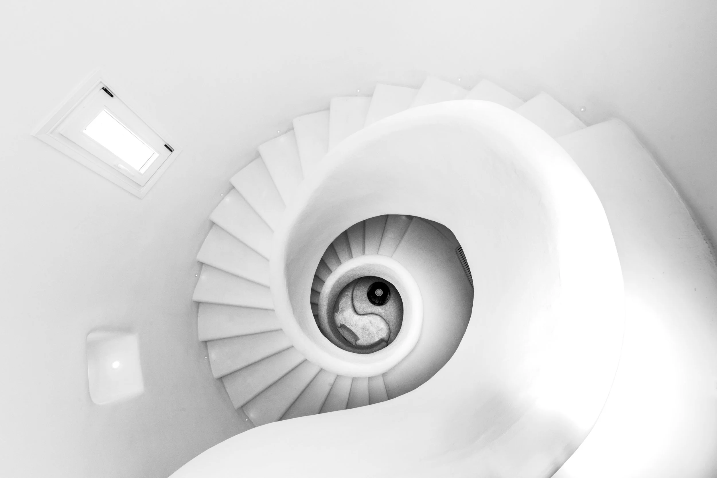 A spiral staircase viewed from above with white steps and walls, featuring a small rectangular window on the wall.