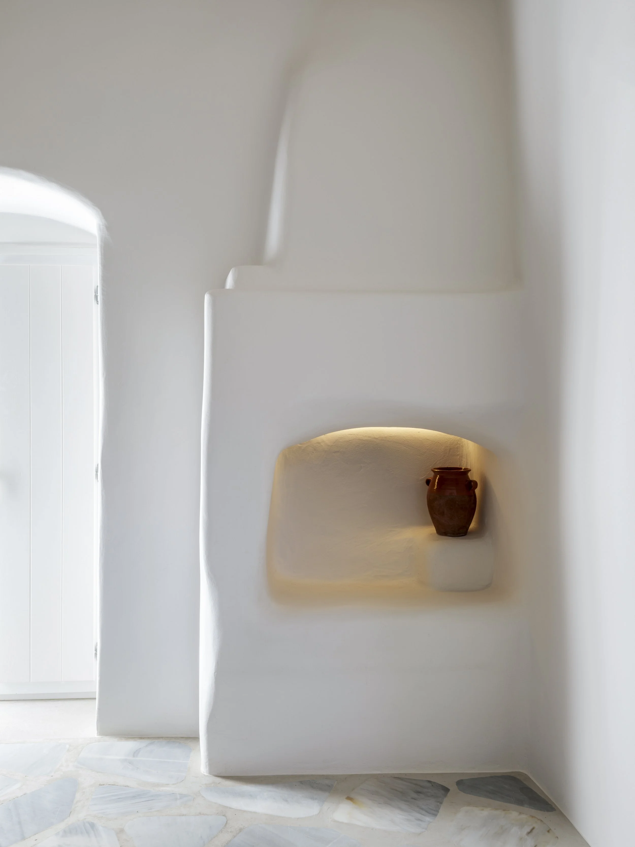 Minimalist white wall with built-in shelves and a small brown vase on one of the shelves, with a natural stone floor visible at the bottom.